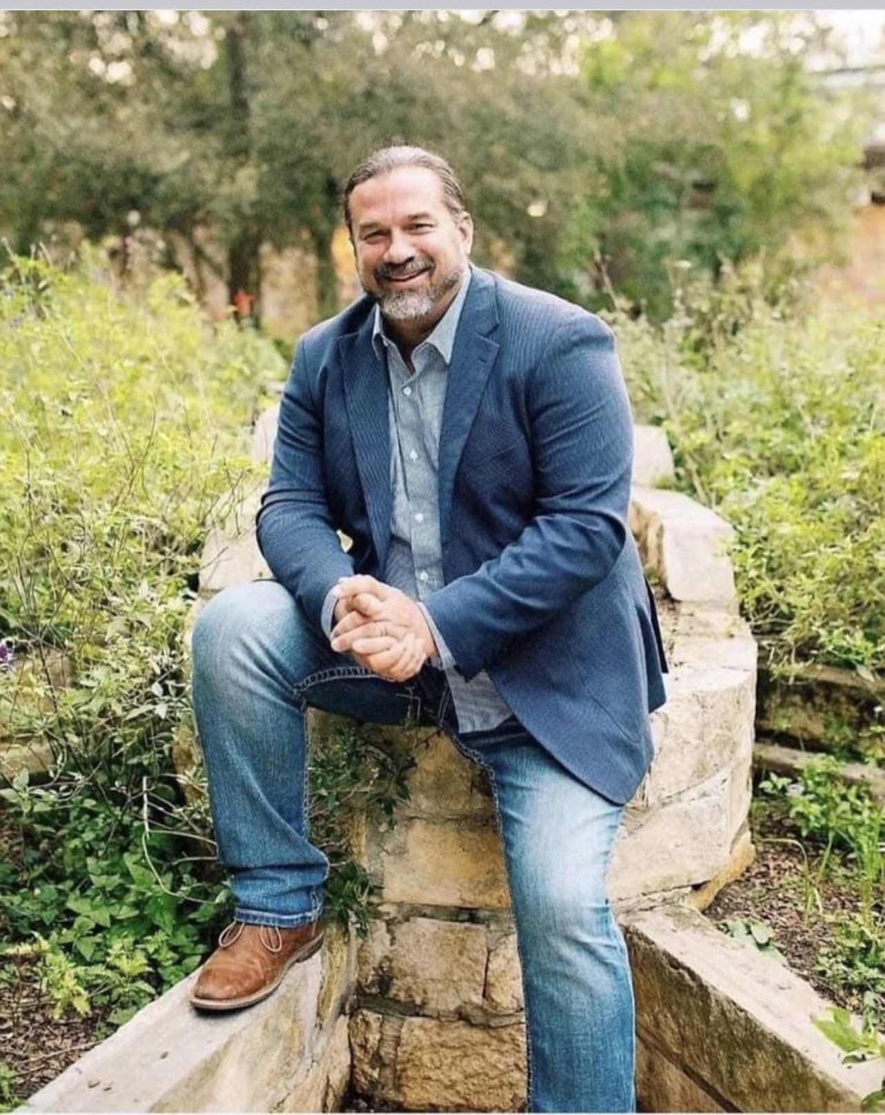 Man in blue blazer and jeans sitting on a stone structure, smiling outdoors.