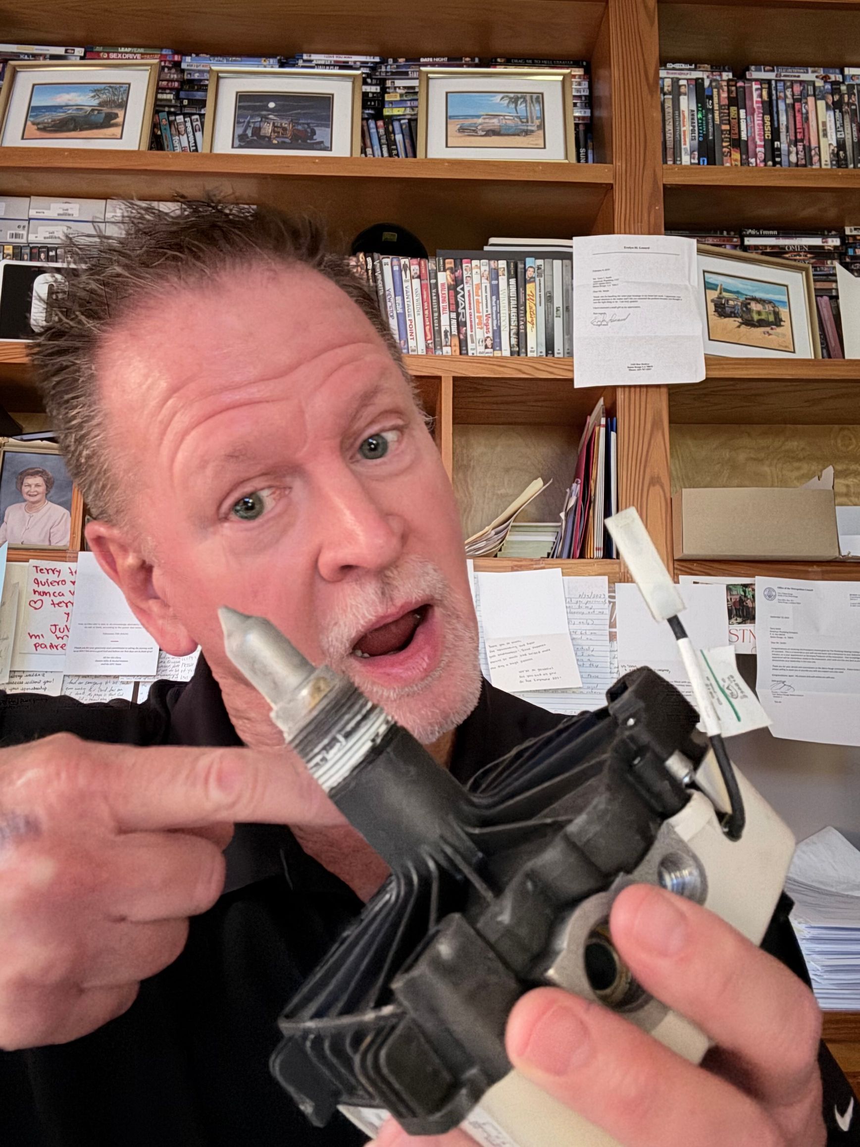 Man holding a large mechanical part up close in a home office, shelves of photos behind him.