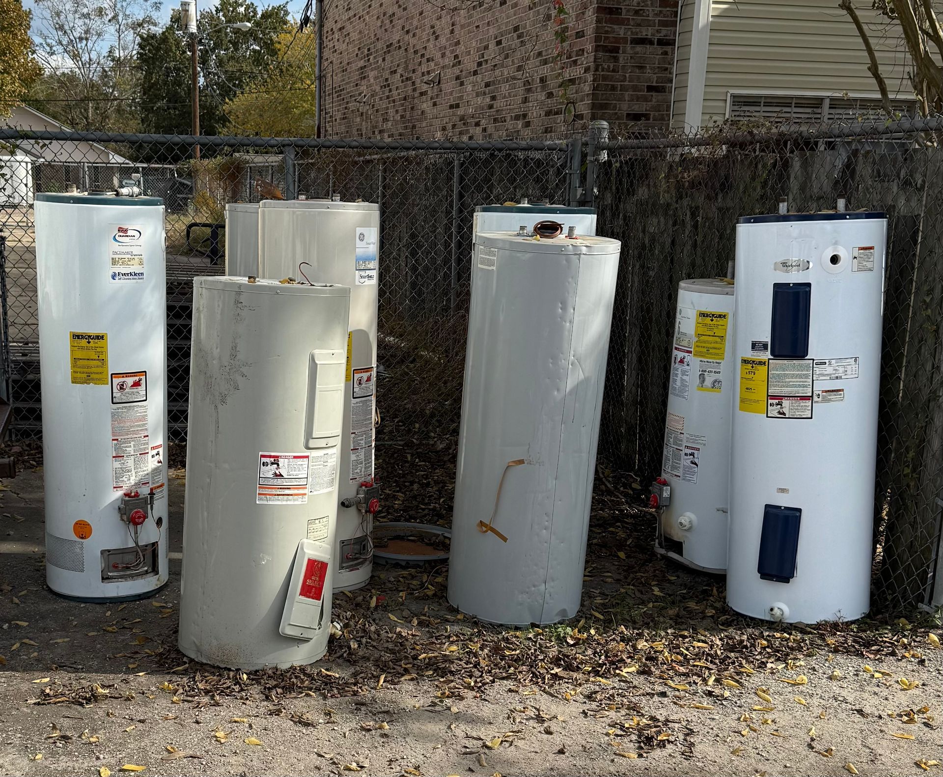 Several large white water heaters standing outdoors on gravel beside a fence.