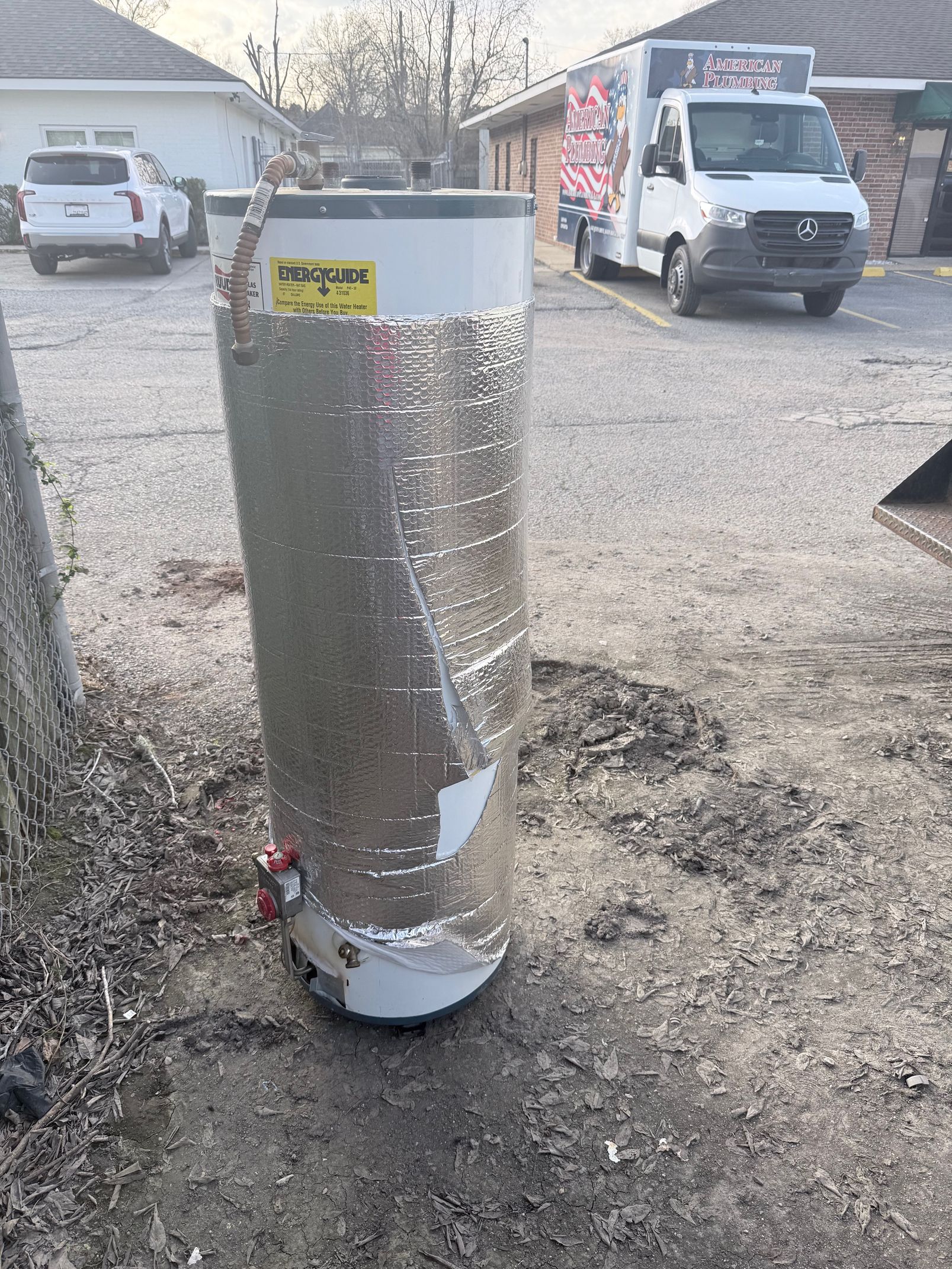 Foil-wrapped insulated cylinder standing in a driveway, with vans and houses in the background.