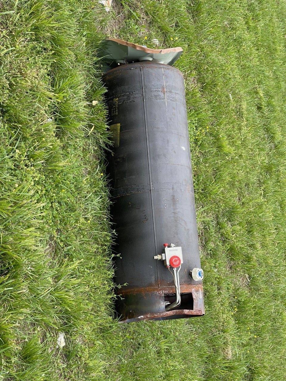 Half of a cylindrical, black water heater lies on green grass. The top is damaged, and a valve is visible.