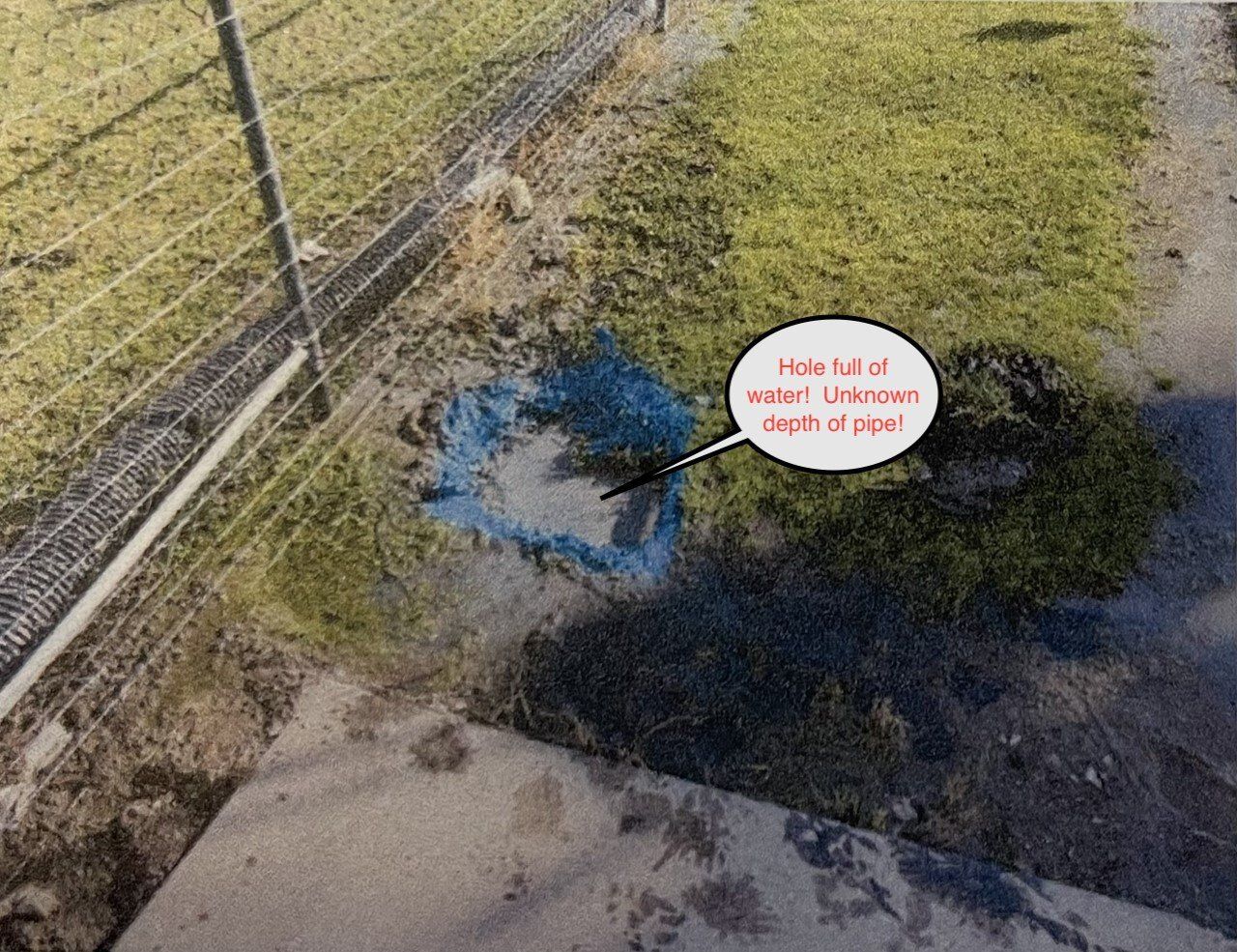 Outdoor grassy drainage area with standing water near a fence and a small sign.