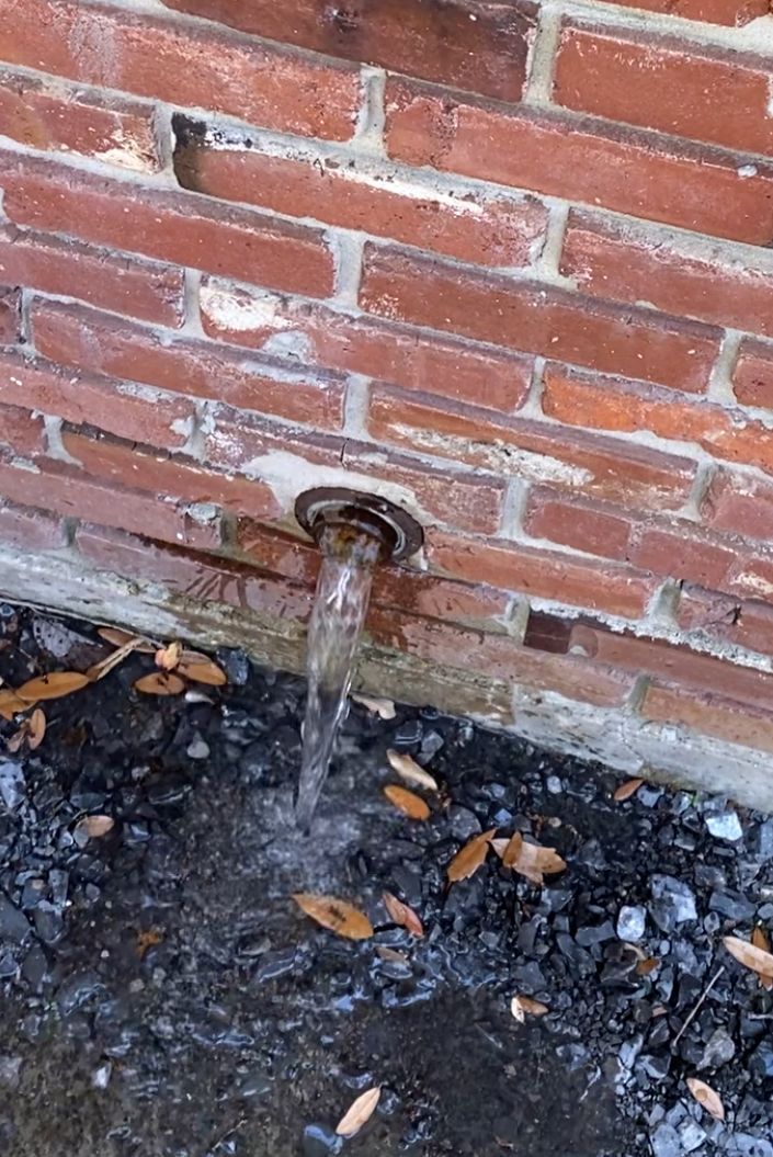 Water flowing from a pipe in a brick wall, onto a gravelly surface.
