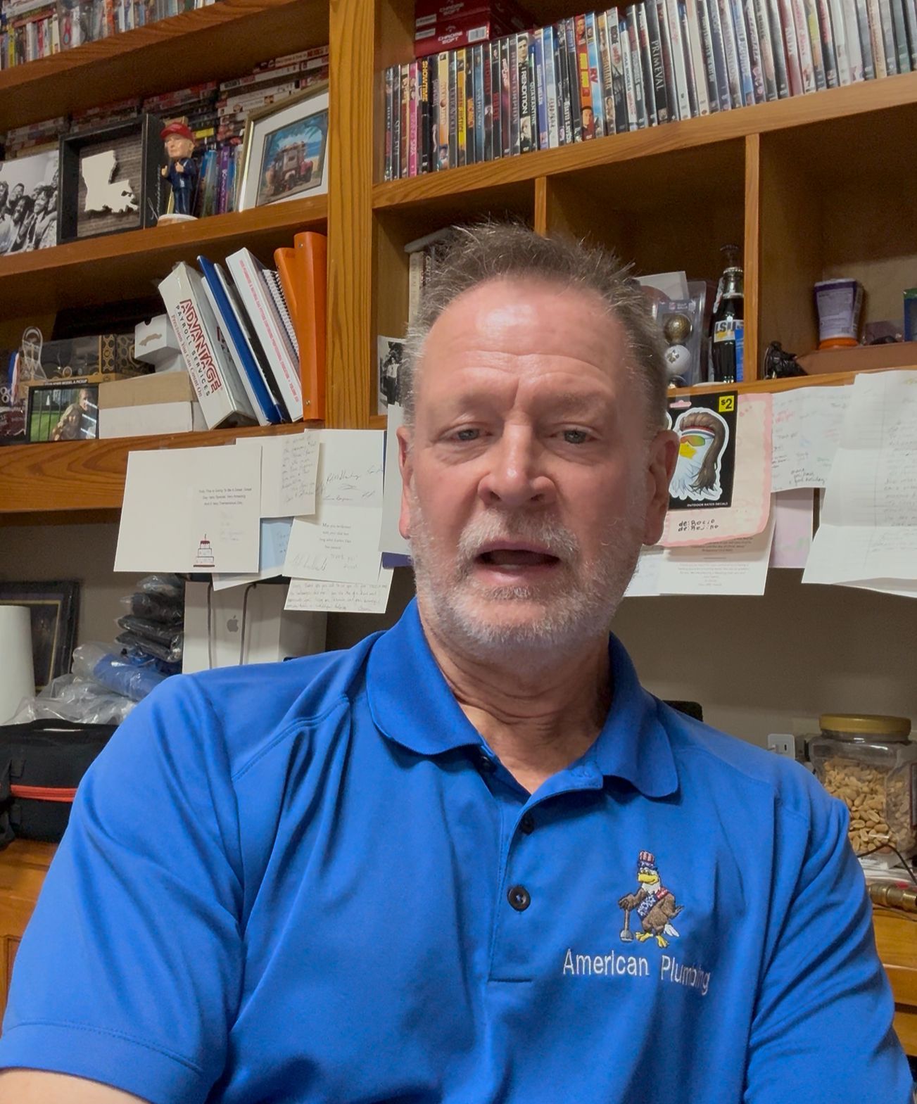 Man in blue polo seated in front of shelves filled with books, DVDs, and papers