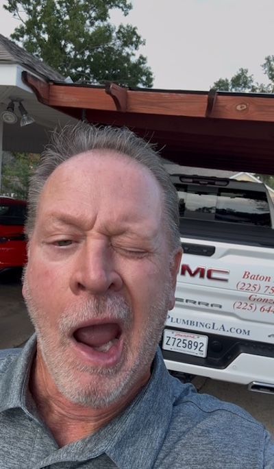 Man winking and yawning in a driveway, with a white GMC truck and carport behind him.