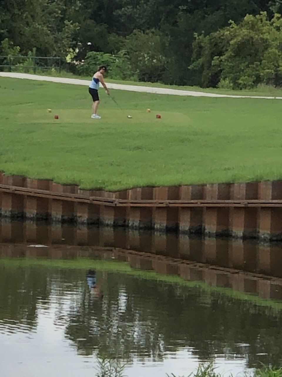 Woman playing croquet on a grassy bank near water, wooden barrier, overcast day.