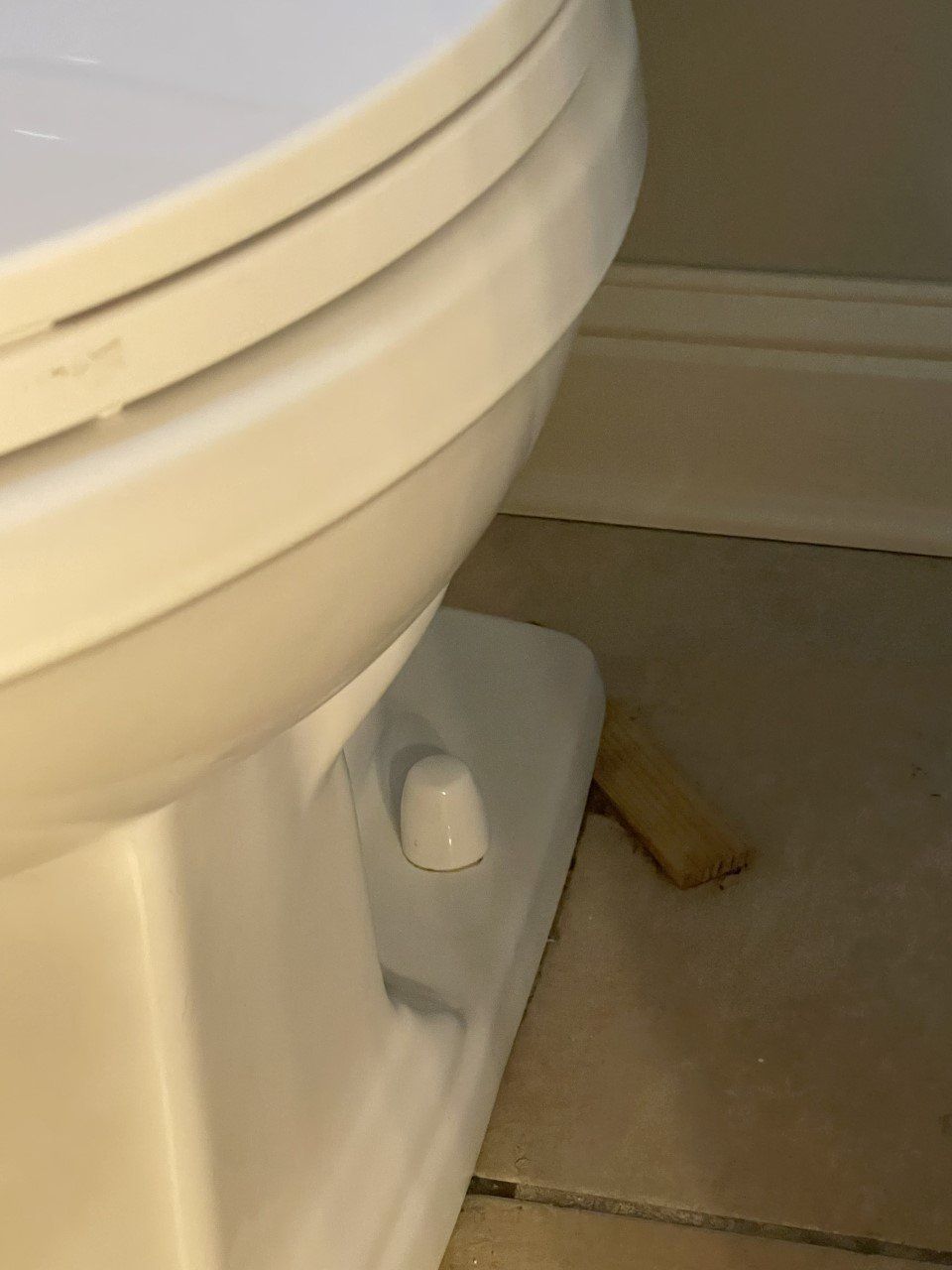 White toilet next to a wall. A small white handle is visible, a piece of wood rests on the tile floor.