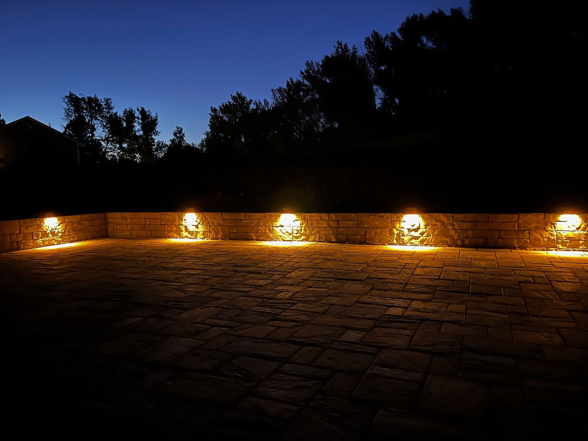 Exterior view of a home at night, with pathway lighting, a lounge chair, and a tree with spotlights.