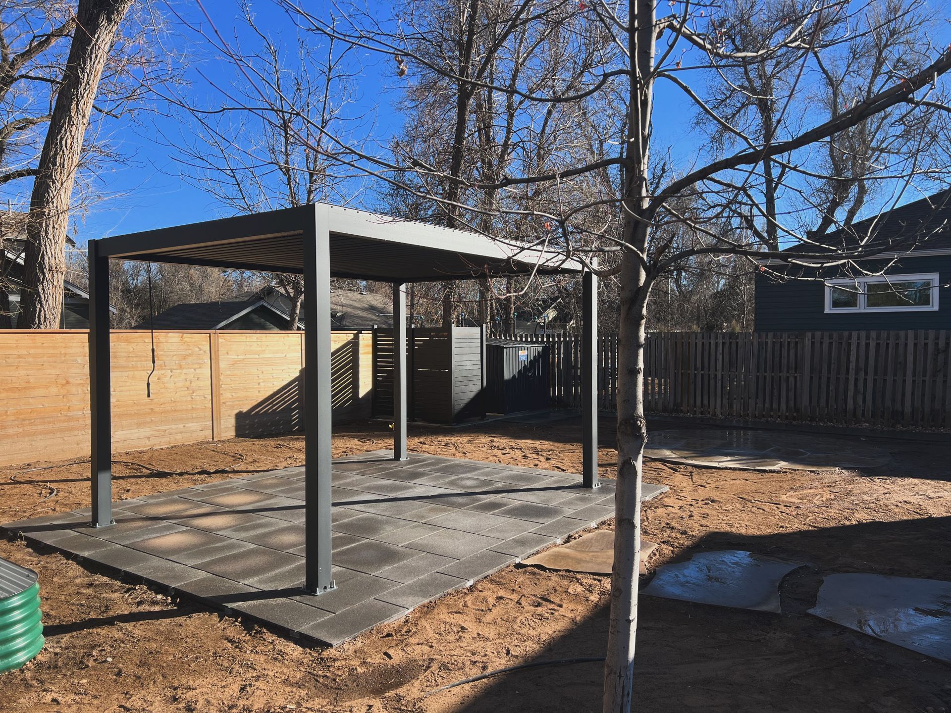 Pergola in a backyard setting; gray structure on a dark gray patio, surrounded by bare trees, wood fence, and brown ground.