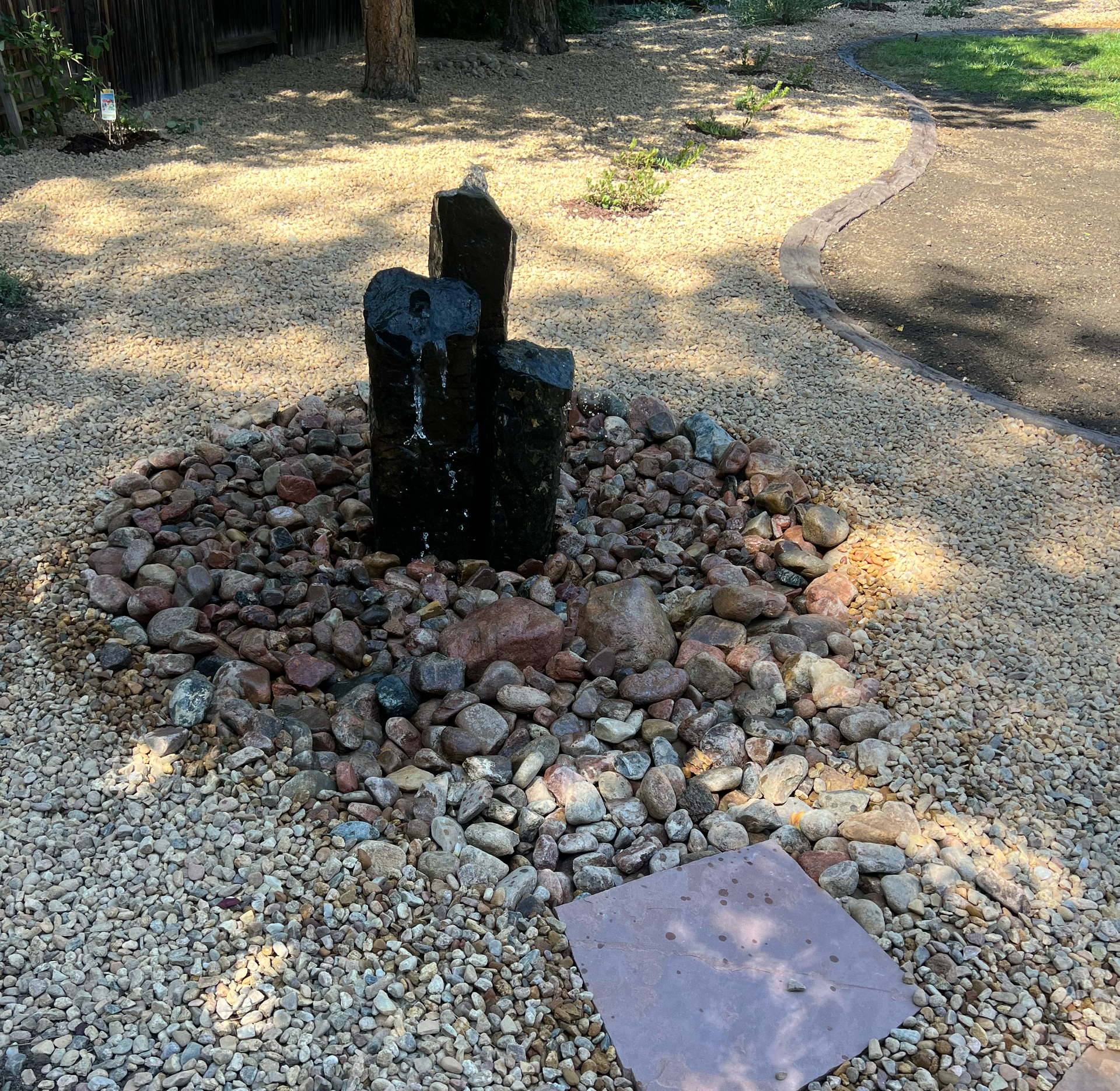 A small, tiered black fountain surrounded by reddish-brown and beige stones. Beige gravel surrounds the feature.