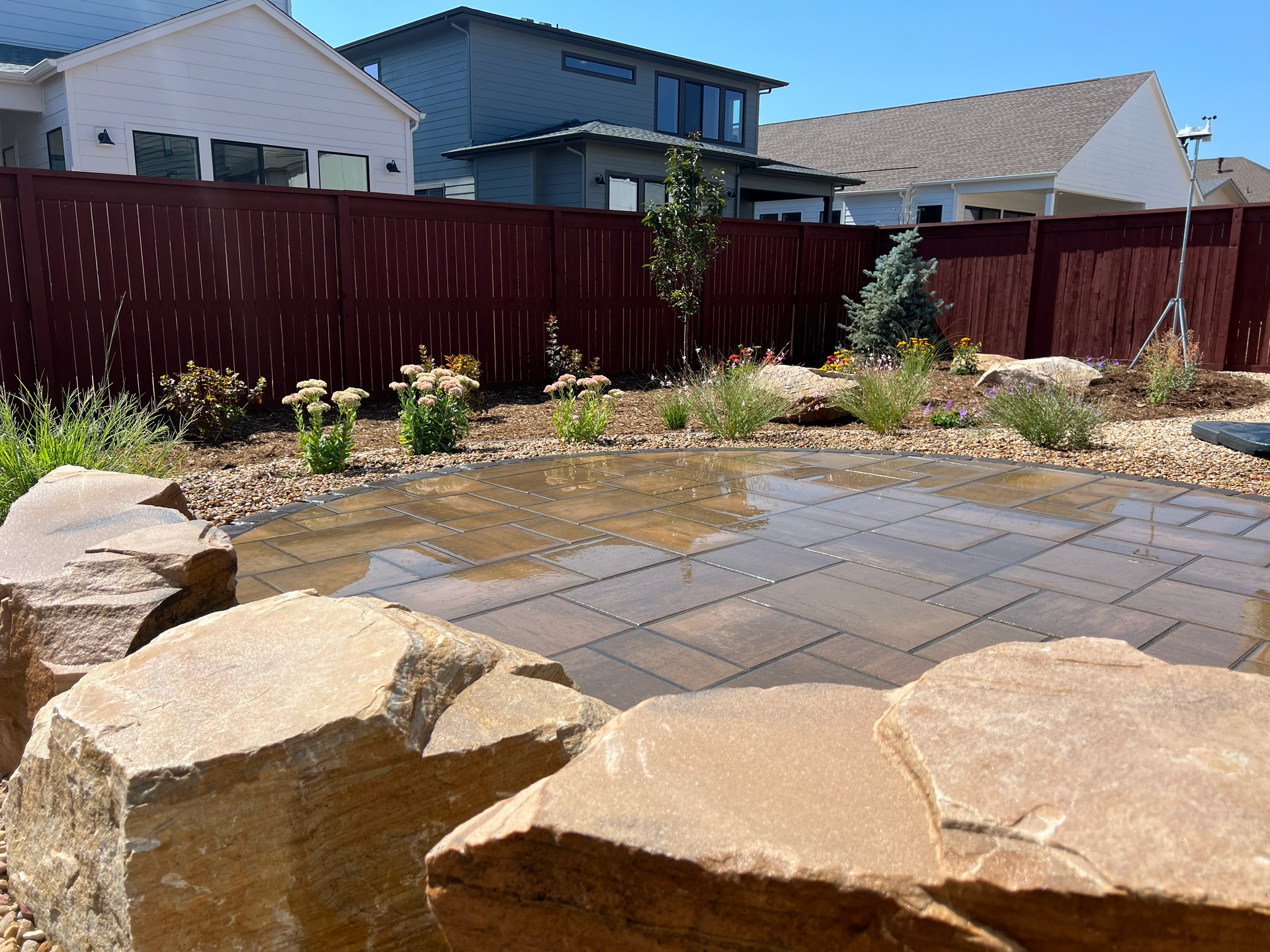 Backyard patio with large rocks, pavers, and a water feature surrounded by plants and a red wooden fence.
