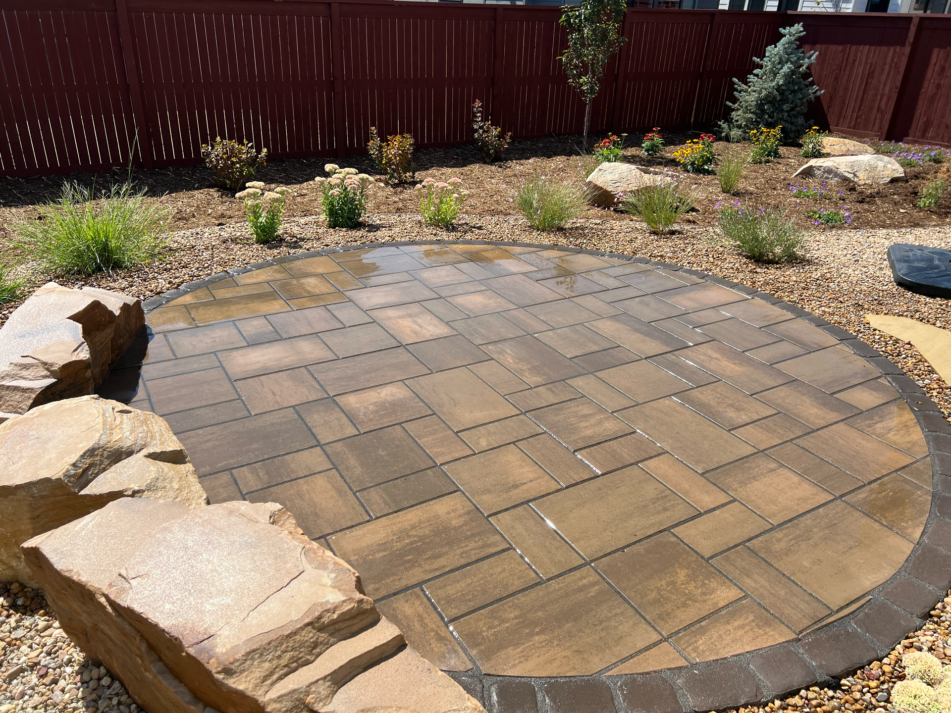 Circular stone patio with rock border surrounded by plants and gravel.