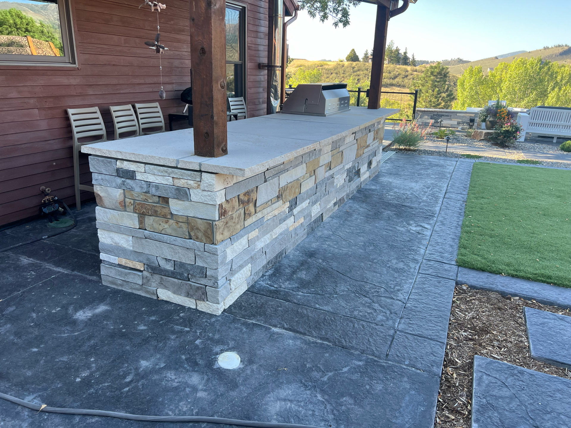 Outdoor stone kitchen island with grill on a stamped concrete patio, next to a lawn.