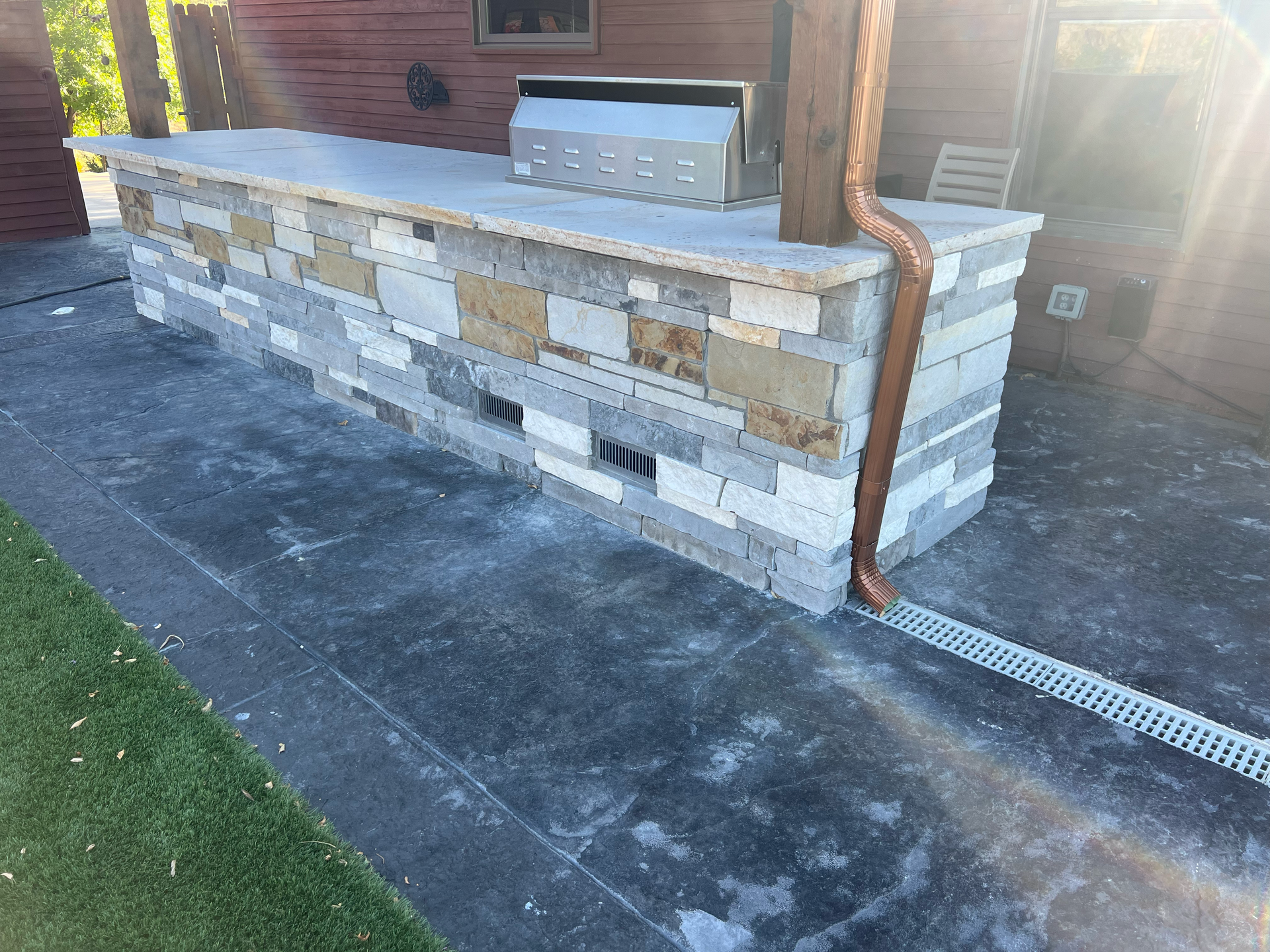 Outdoor stone-faced kitchen with grill and concrete countertop, on a patio next to grass.
