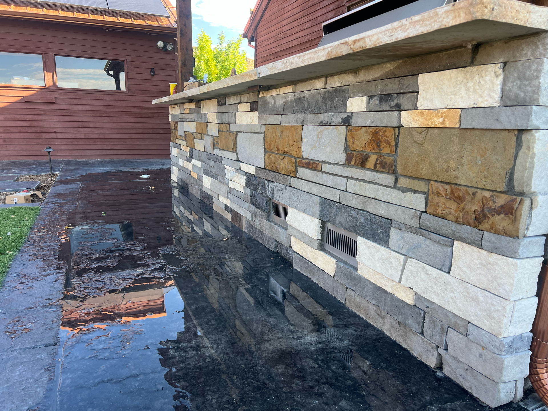 Stone-clad outdoor kitchen counter with varying colored rectangular stones, countertop, and wet black tiled floor.