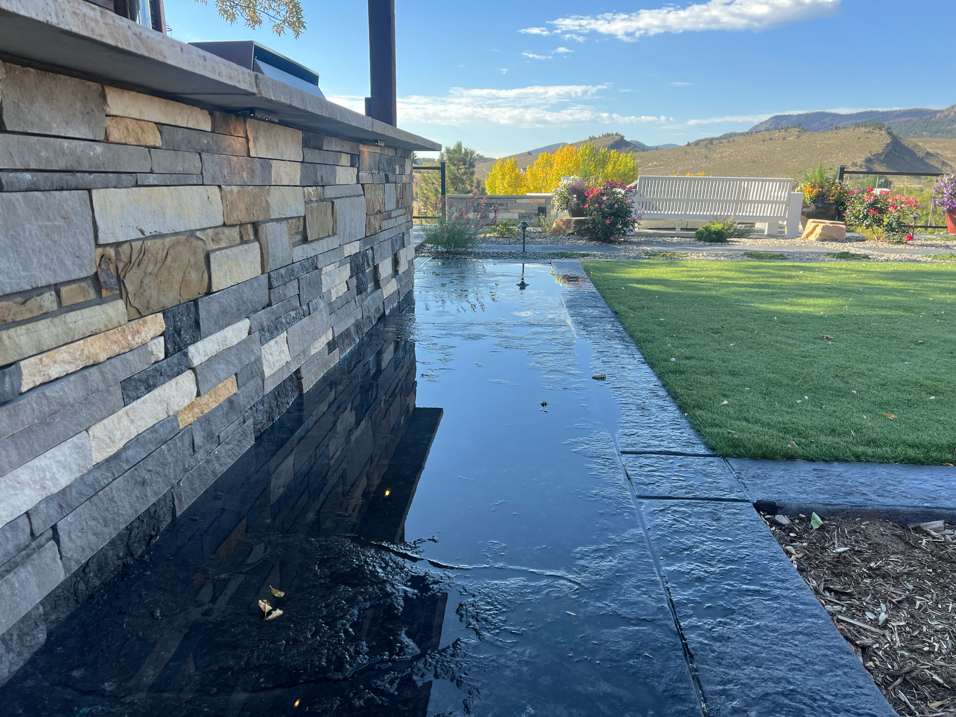 Puddled water along a dark stone border next to a stone wall and green lawn, with a distant outdoor seating area.