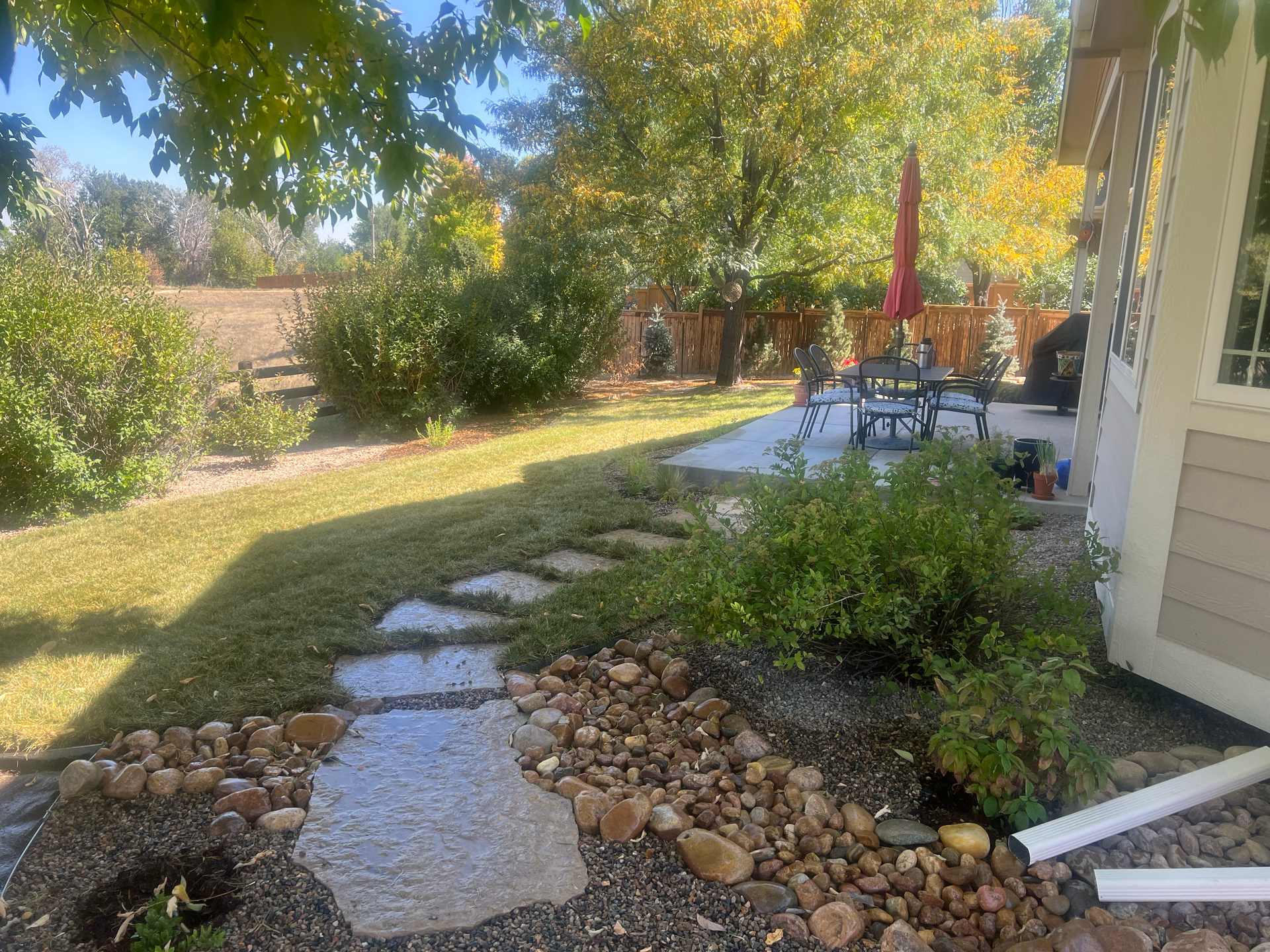 Stone path leads through a yard with rocks, grass, and a patio with table and umbrella.