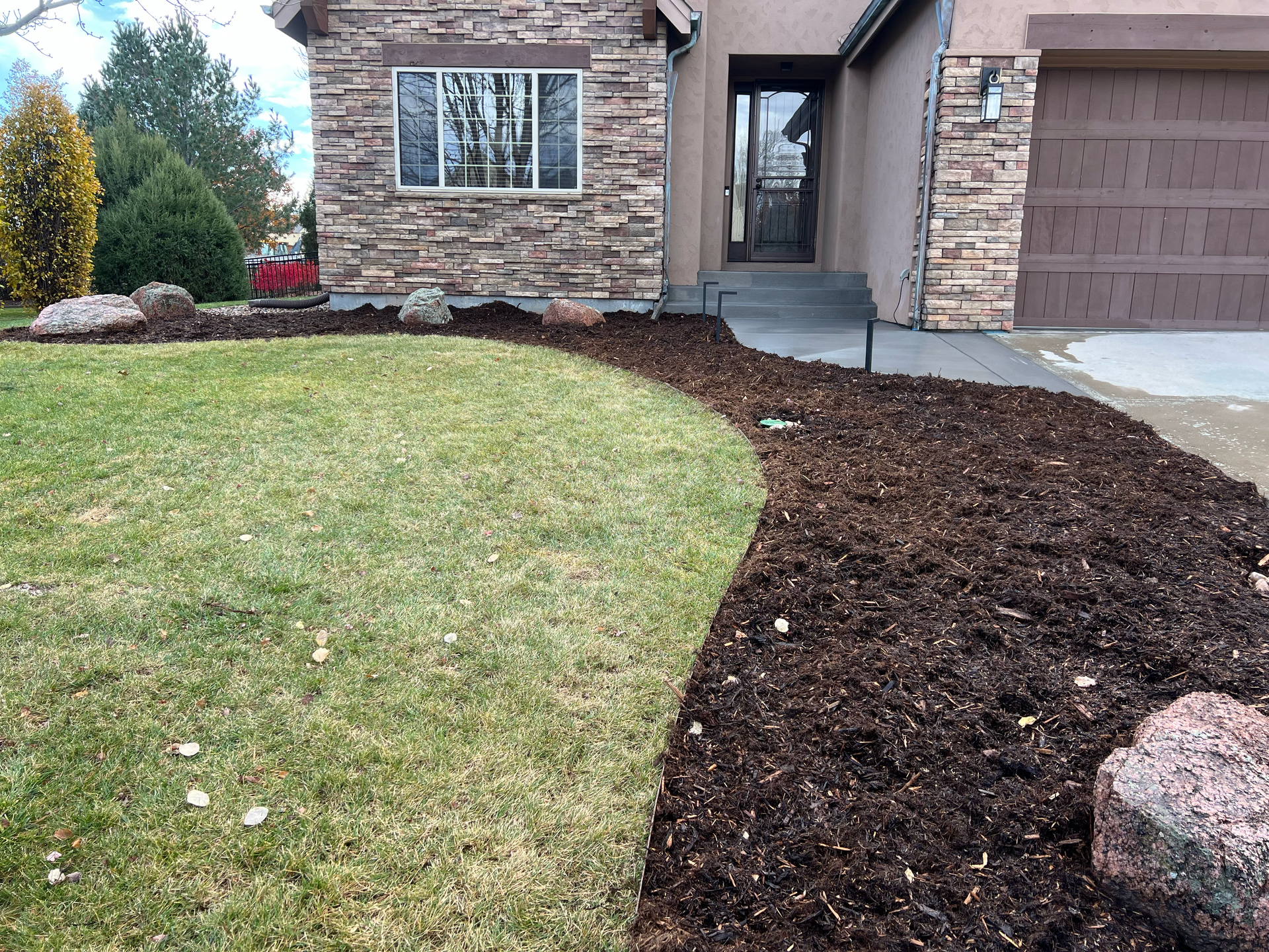 Lawn with a mulch border leading to a house with stone siding and a brown garage door.