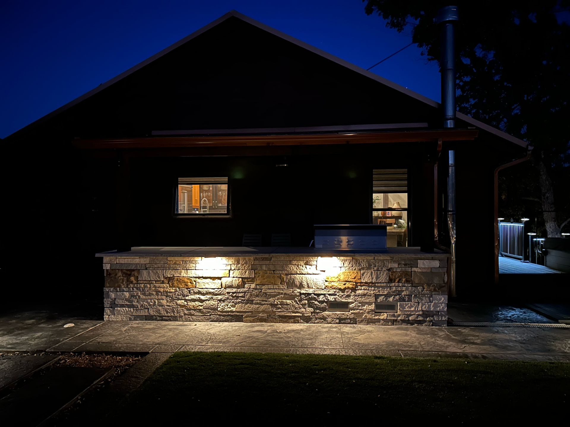 Nighttime shot of a brick outdoor kitchen with lit accent lights, beneath a dark house with lit windows.