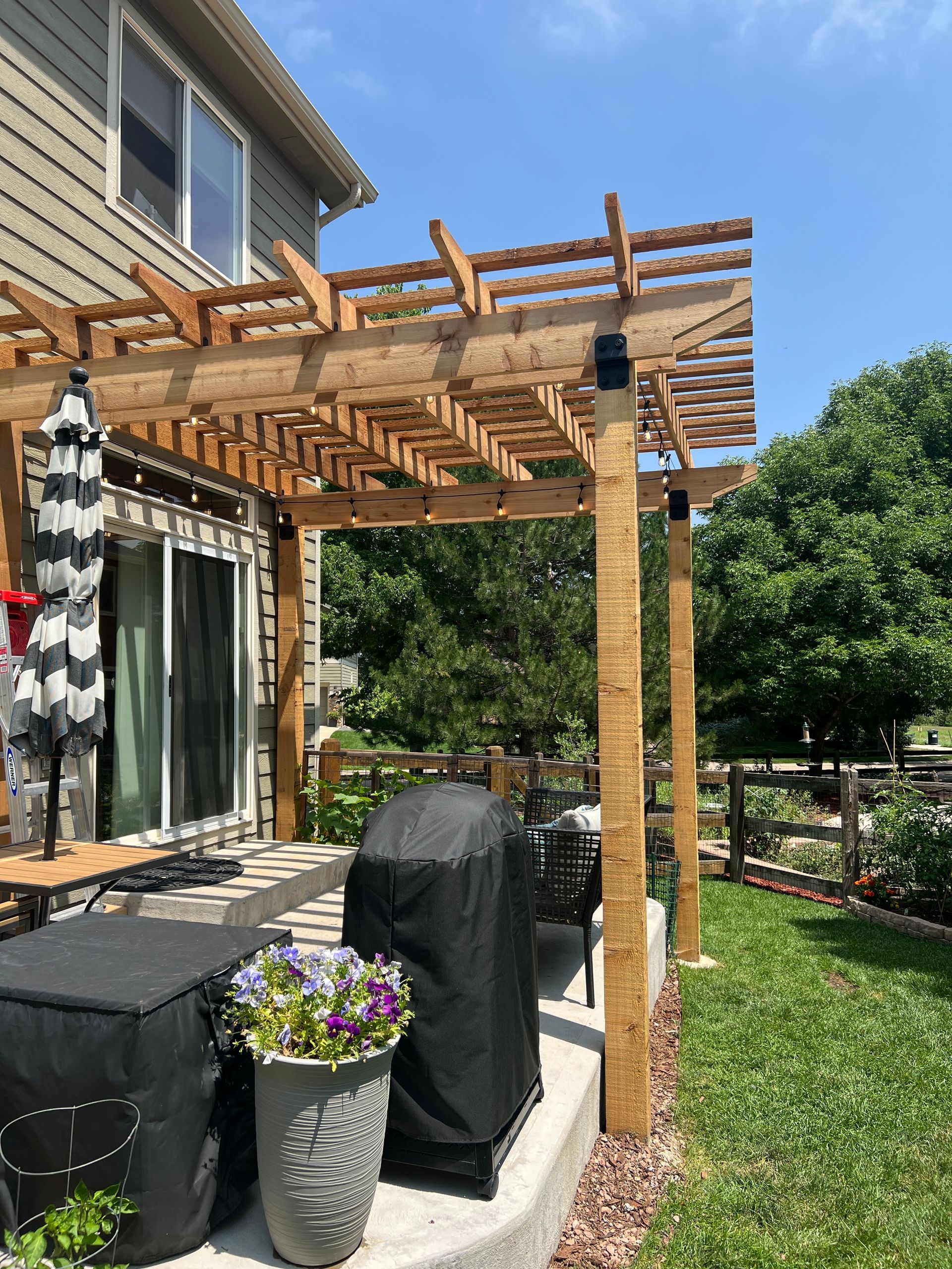 Wooden pergola over a patio with a grill, table, and potted flowers.  Backyard setting with grass and trees.