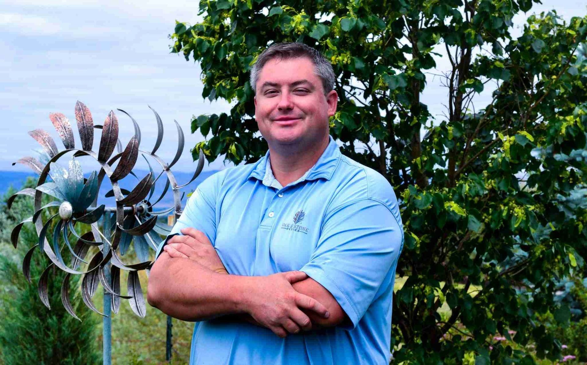 Man in blue polo shirt with arms crossed, standing near a metal pinwheel and greenery.