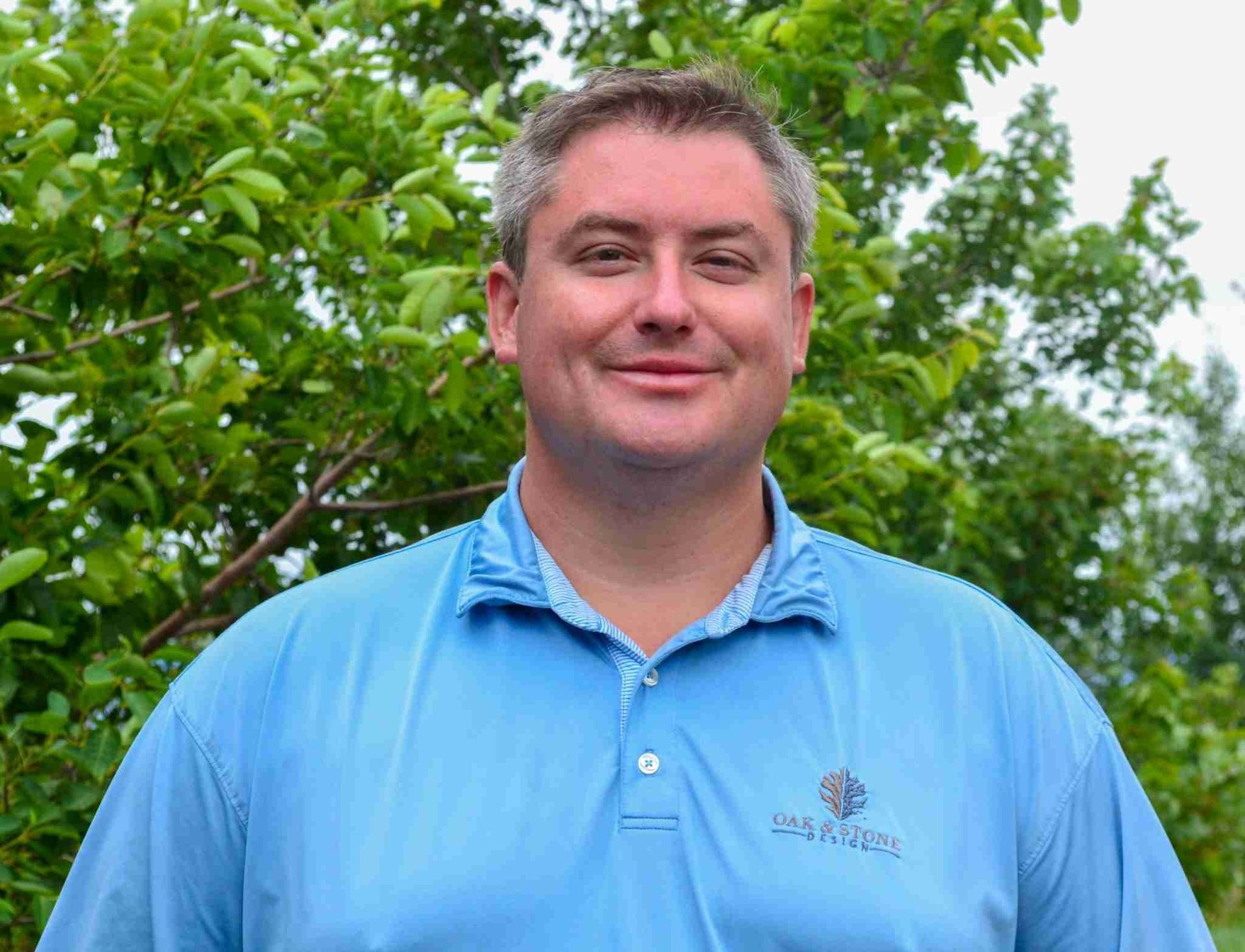 Man in blue golf shirt smiling outdoors, with green foliage background.