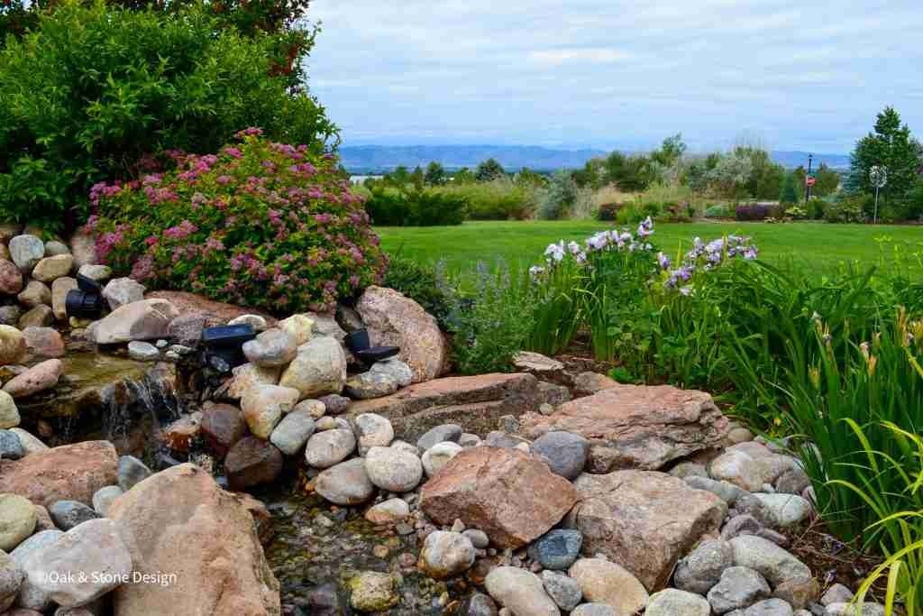 Rocky water feature with cascading water, surrounded by green grass and plants. Cloudy sky.