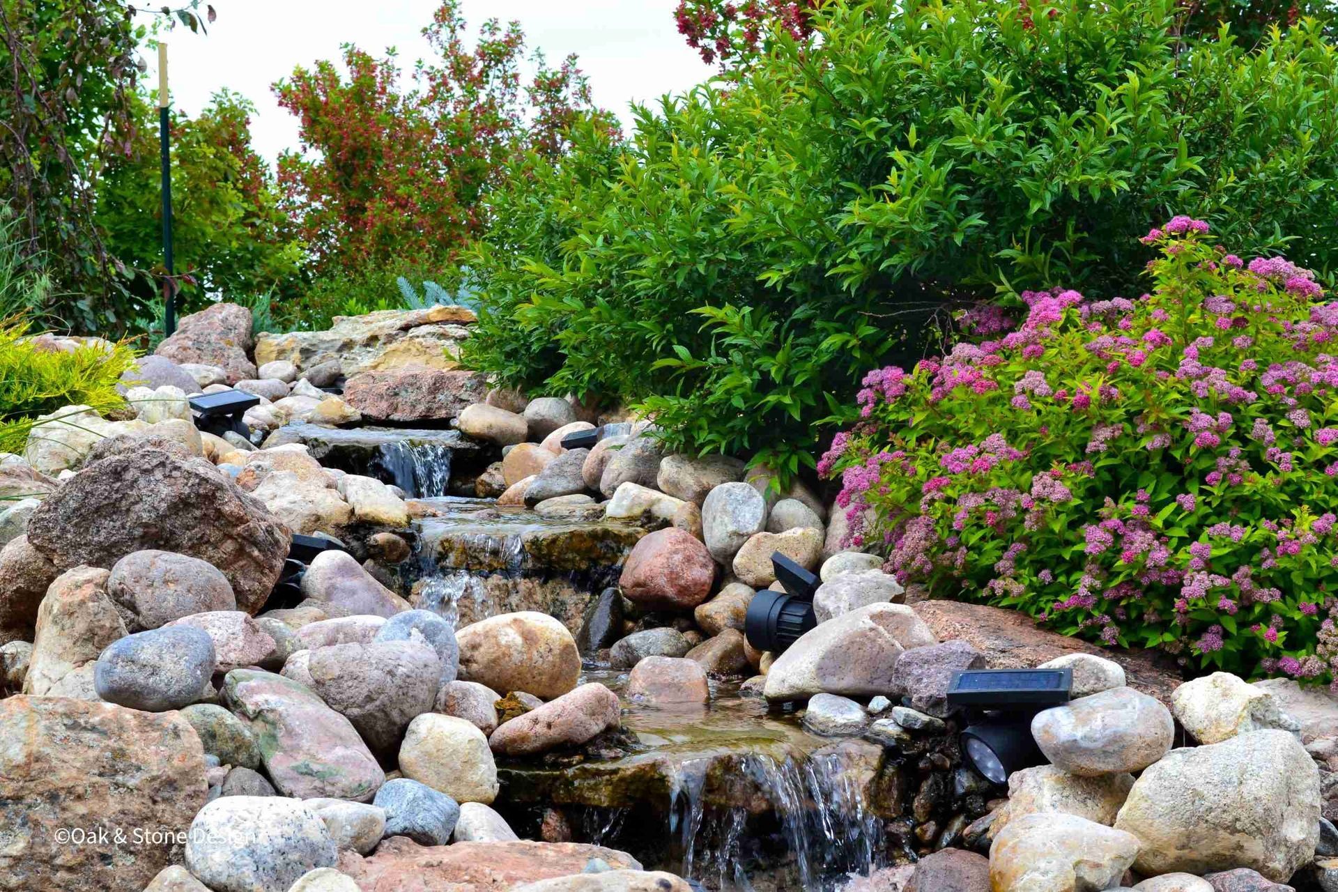 Water cascading down rocky steps, surrounded by green shrubs and pink flowers.