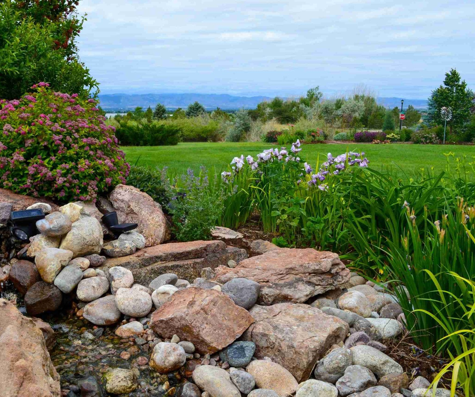 Stone patio with seating and umbrella near a house, surrounded by landscaping on a sunny day.