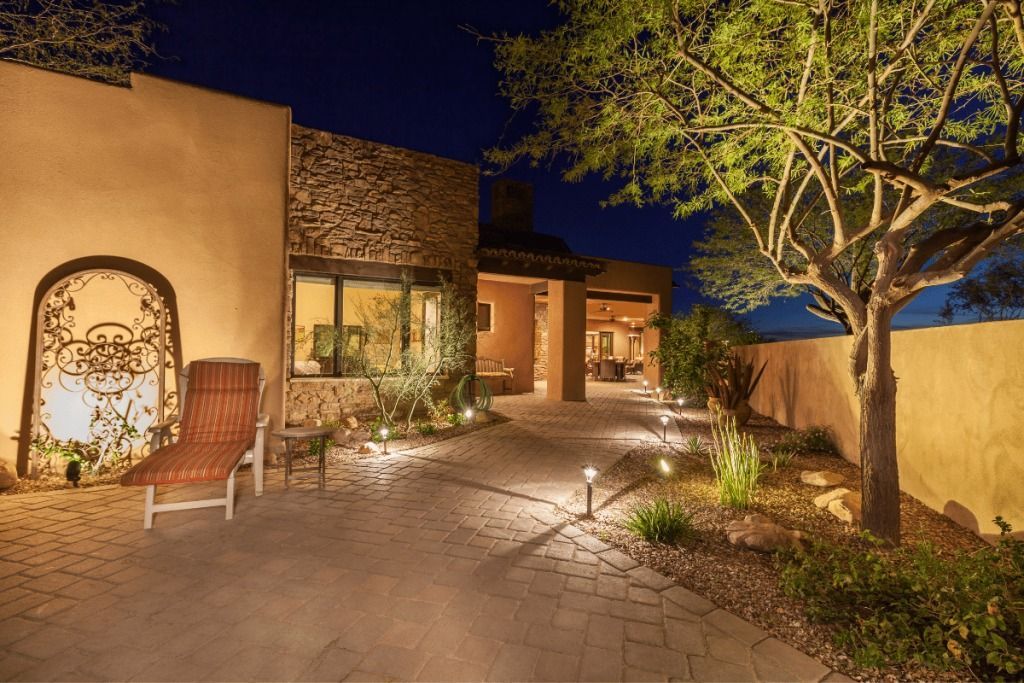 Exterior view of a home at night, with pathway lighting, a lounge chair, and a tree with spotlights.