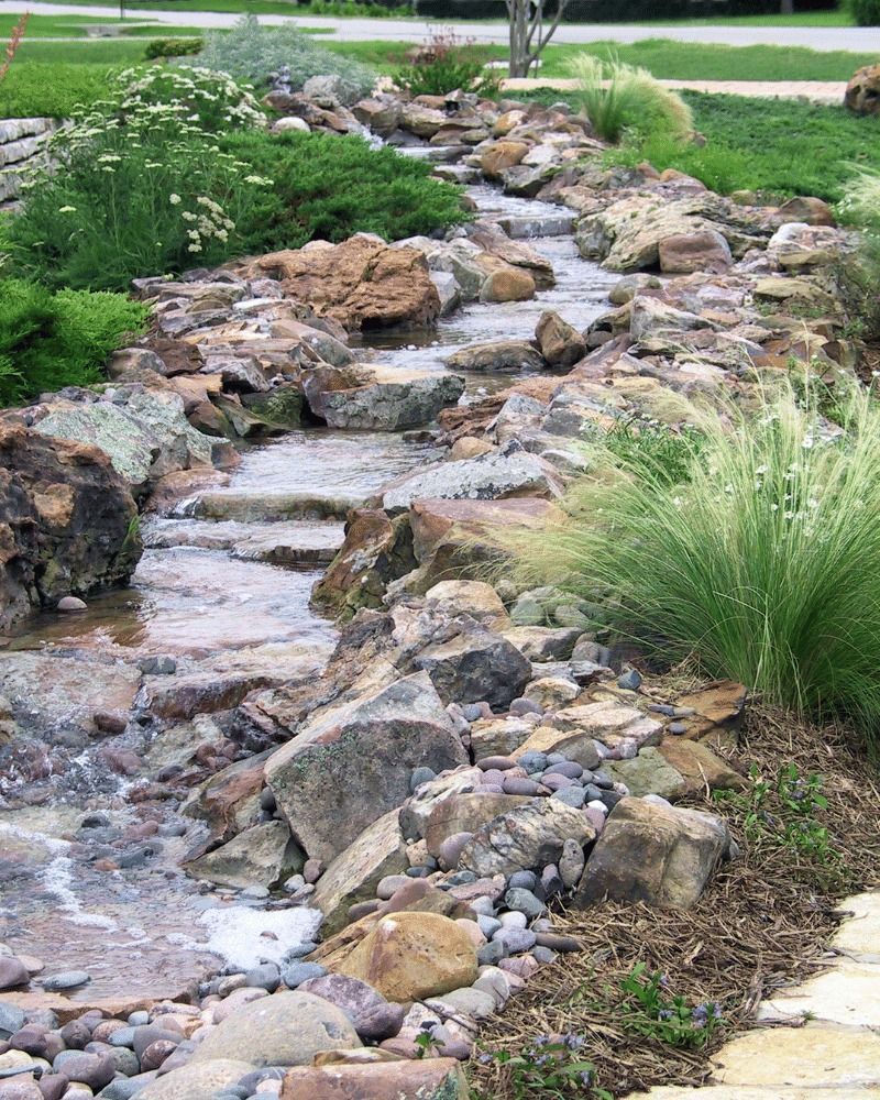 A small stream flows through a rock garden with greenery alongside.