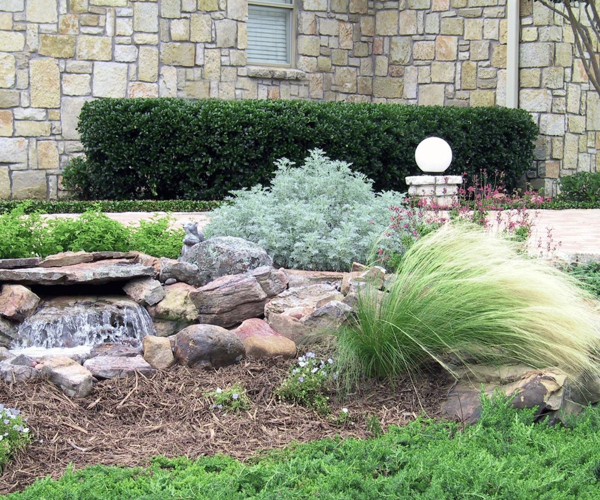 A garden with a small waterfall, various plants, and a round light fixture in front of a stone wall.