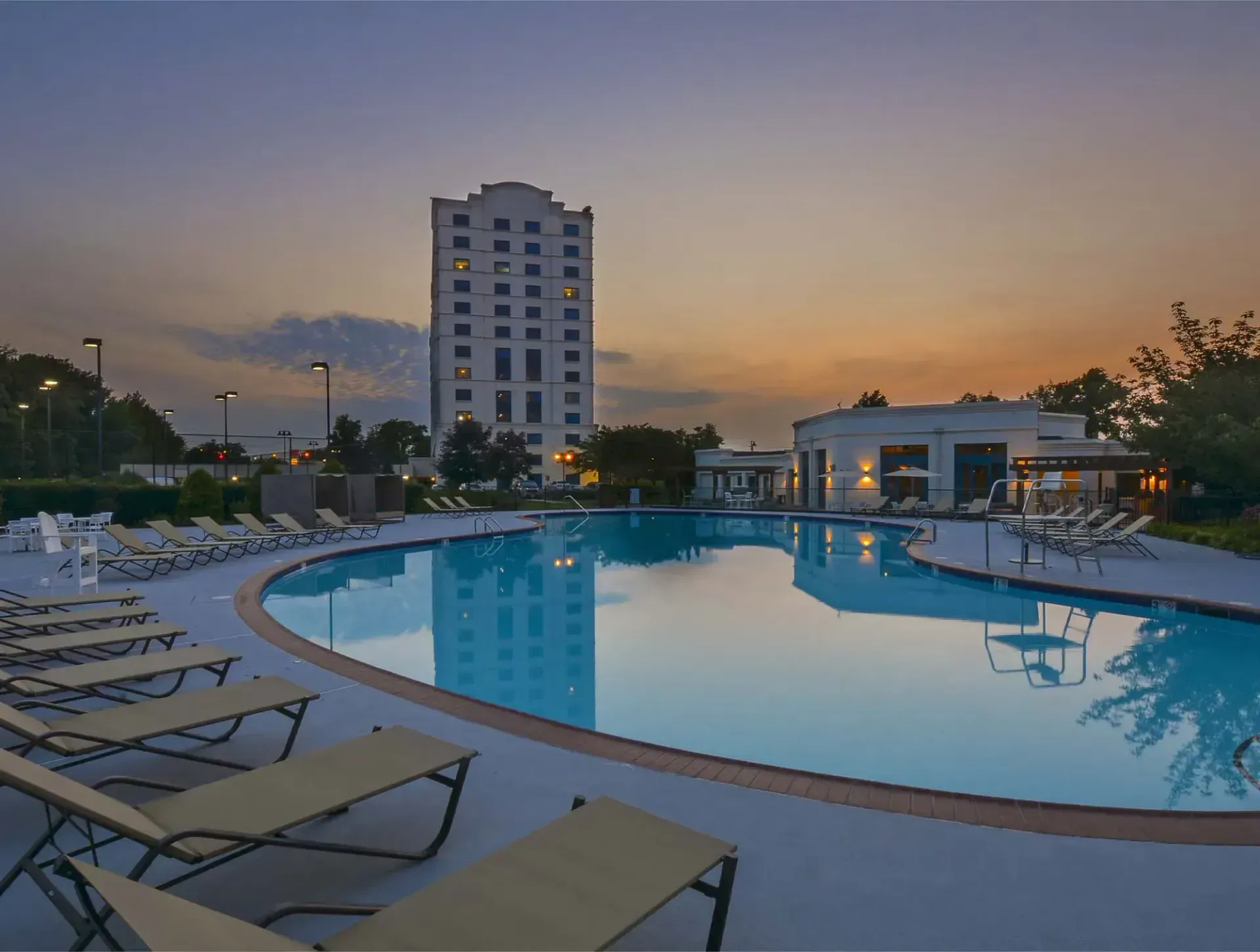 Swimming pool in front of a tall white building at dusk. Lounge chairs surround the pool, under a colorful sky.