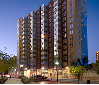 Multi-story apartment building with tan, blue, and silver facade. Street-level shops and streetlights. Evening light.