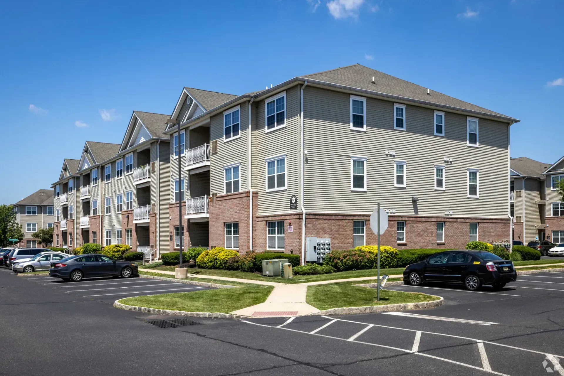 Apartment complex exterior with beige siding, brick accents, and parked cars on a sunny day