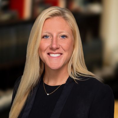 Blonde woman smiling, wearing a black blazer and necklace. Blurred office backdrop.