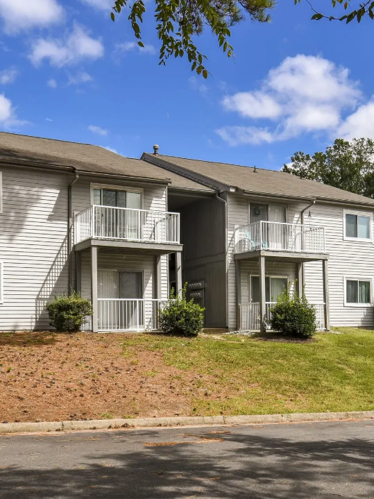 Two-story apartment building with white siding, small balconies, and a grassy front slope under a blue sky