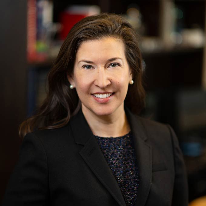 Woman with brown hair smiles, wearing a black blazer over a patterned top, in an office setting.