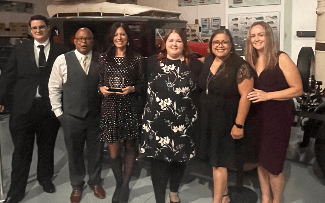 Group of six people posing with an award, standing in front of vintage vehicles.
