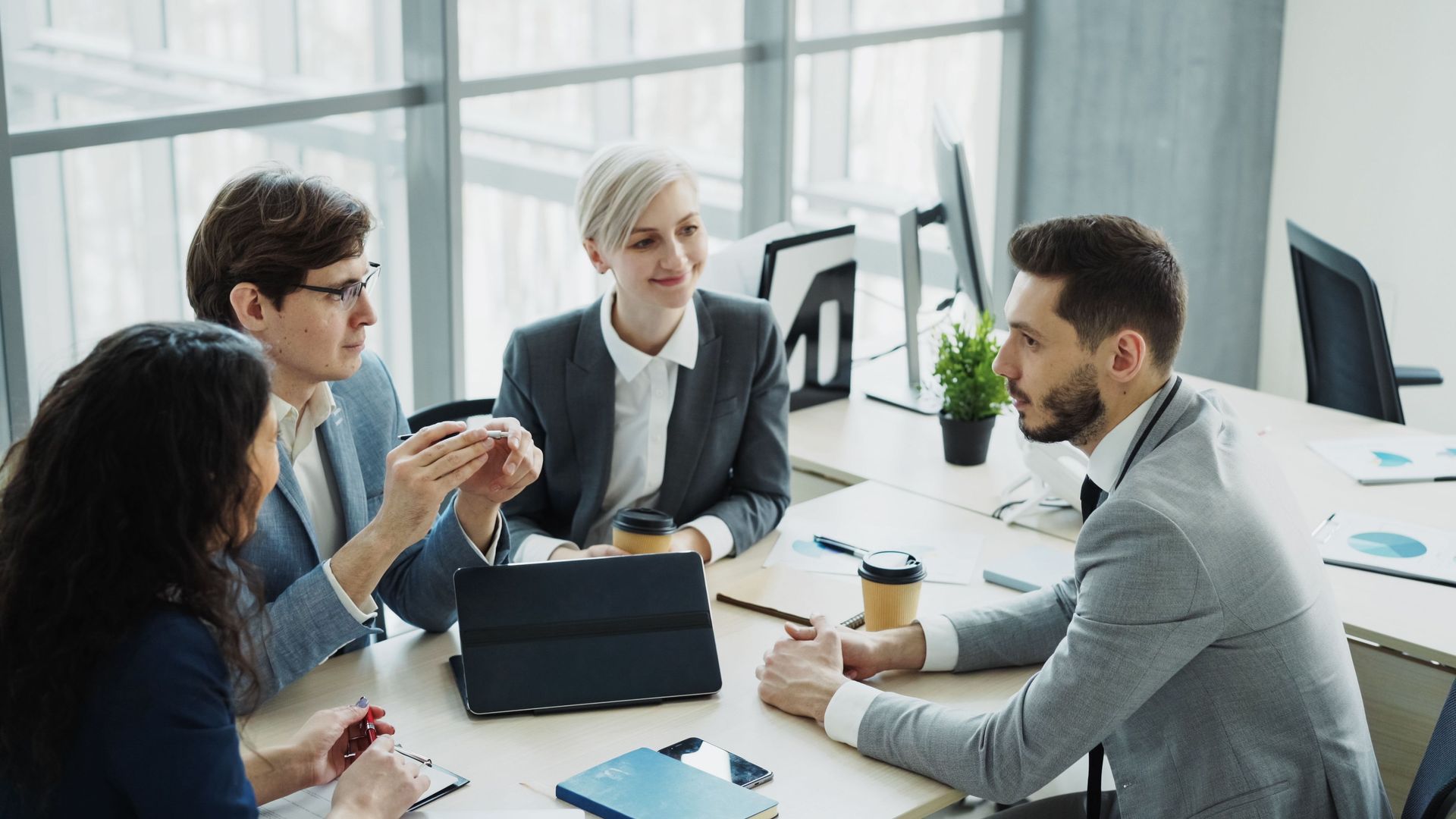 Four coworkers in a bright office discussing around a laptop with coffee cups on the table