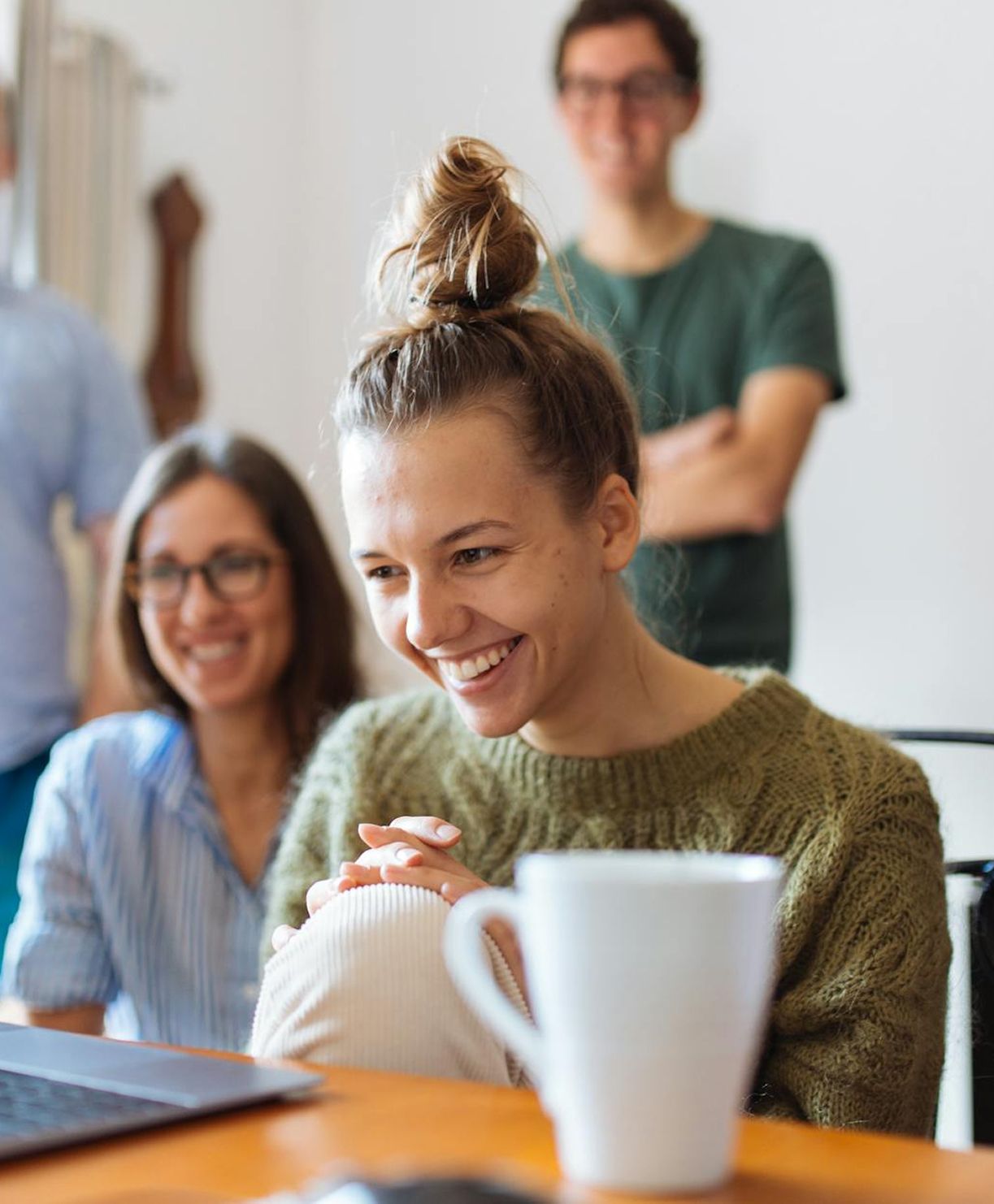 Woman with bun smiles, looking at laptop; colleagues in background. Cup on desk.