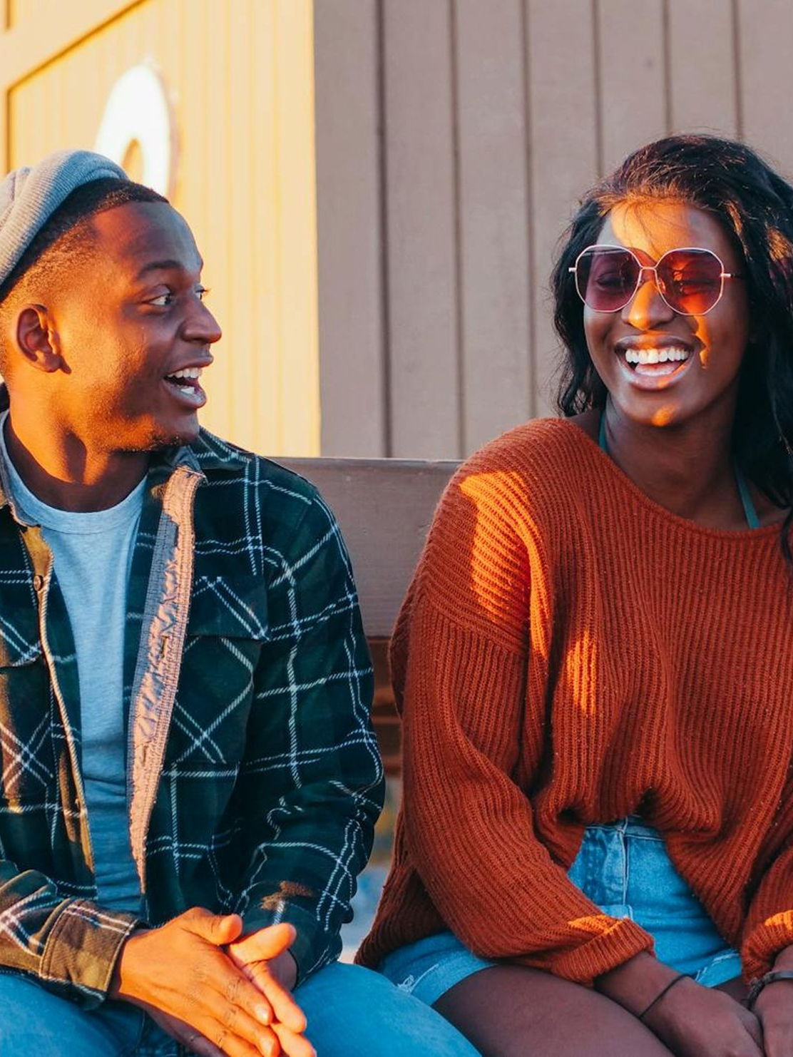 Two people smiling, outdoors. Man in plaid shirt, woman in sunglasses and sweater.