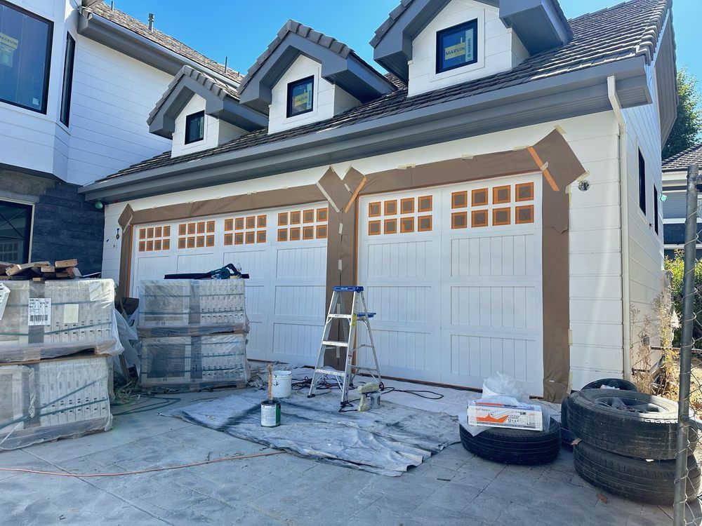 Two-car garage exterior being painted; white doors, tan trim. Bricks and painting supplies are visible.