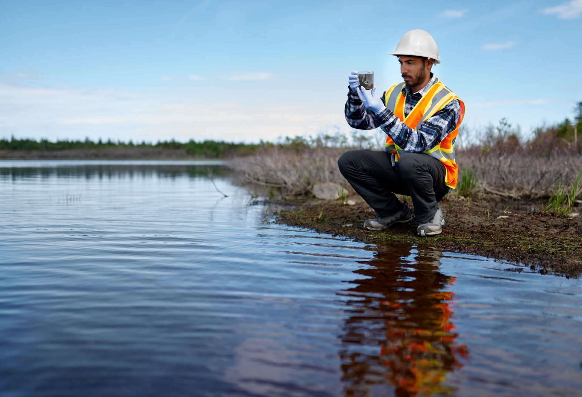 Worker in hard hat and safety vest taking a water sample beside a river