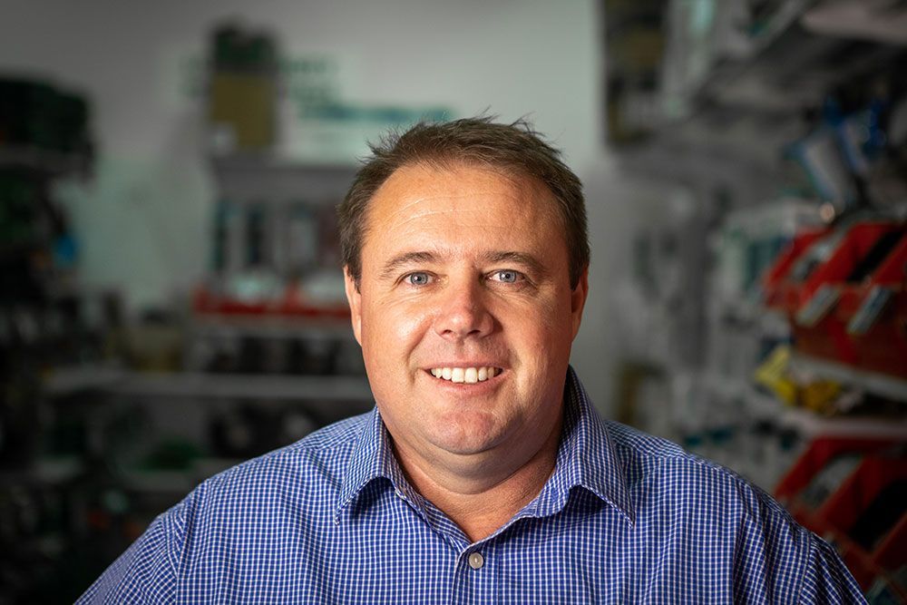 Smiling man in a blue checkered shirt standing in a workshop, with blurred tools in the background