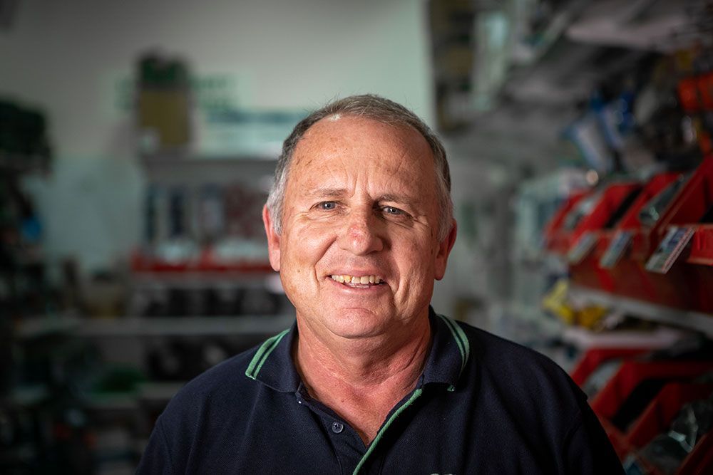 Man smiling in a workshop, wearing a dark polo shirt, with tools and equipment blurred in the background.