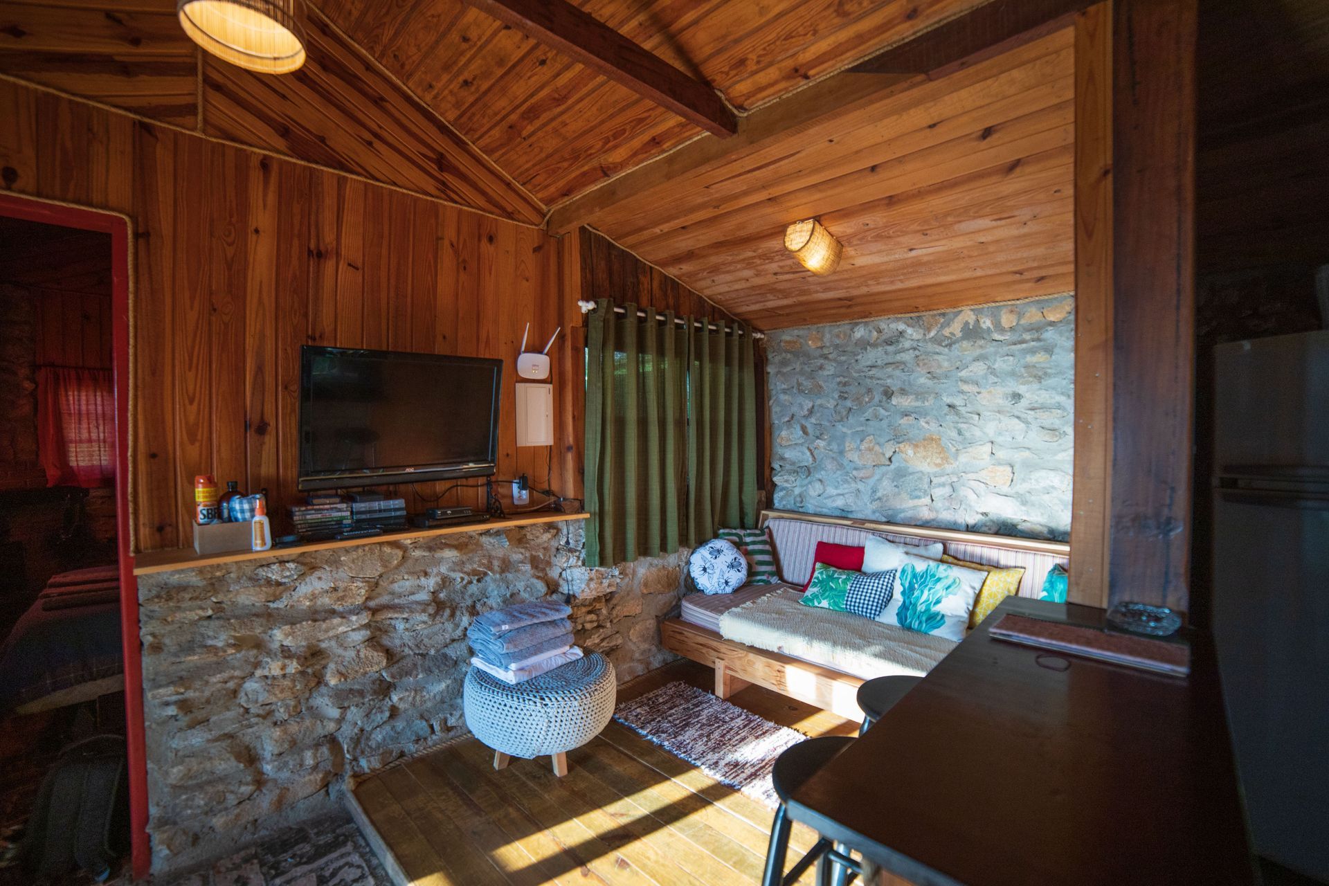 Rustic wooden cabin interior with stone fireplace, sofa, woven chair, and sunlight from windows