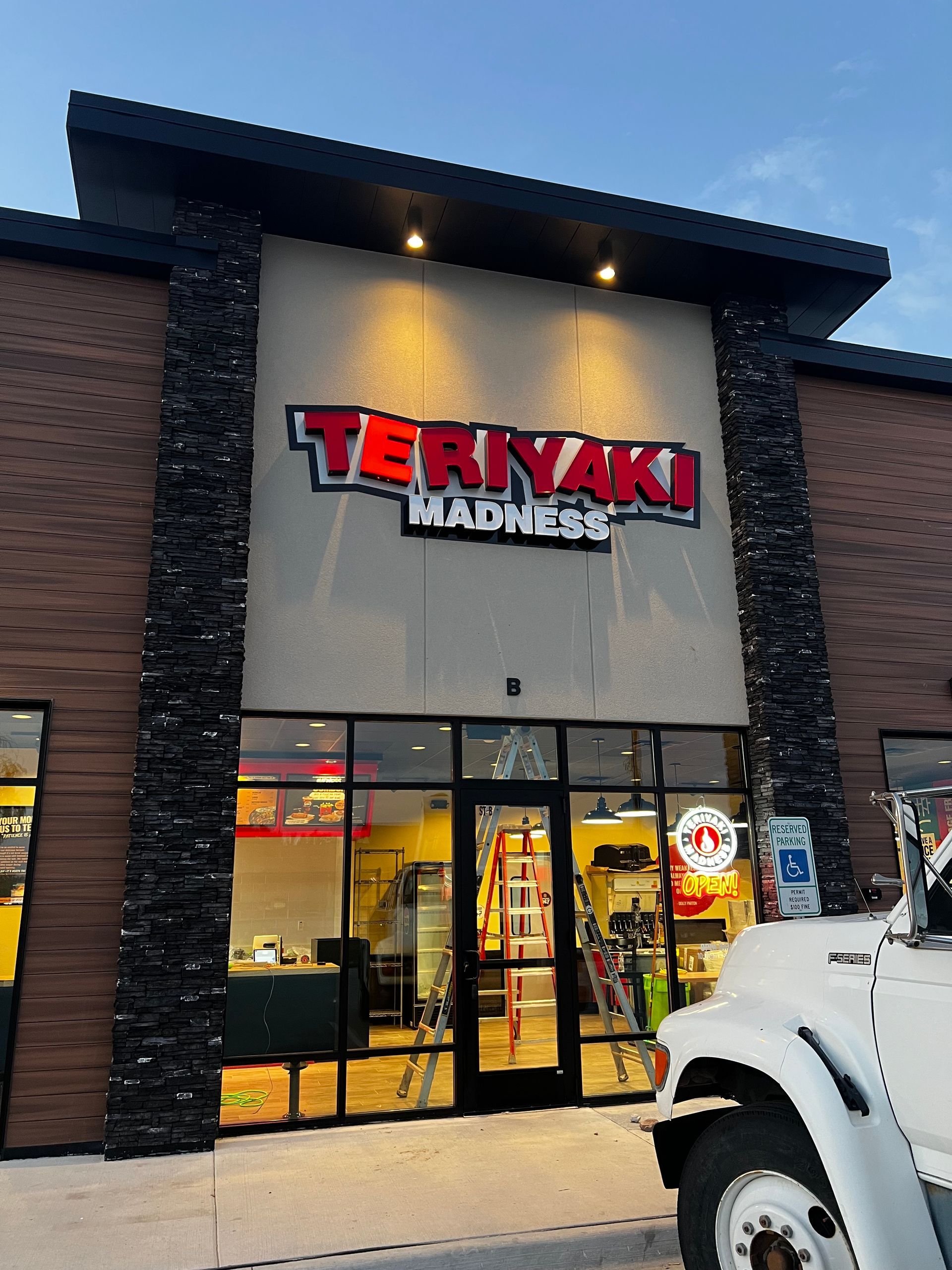 Teriyaki Madness restaurant exterior with illuminated red sign. Black accents, glass doors, and a white vehicle are visible.