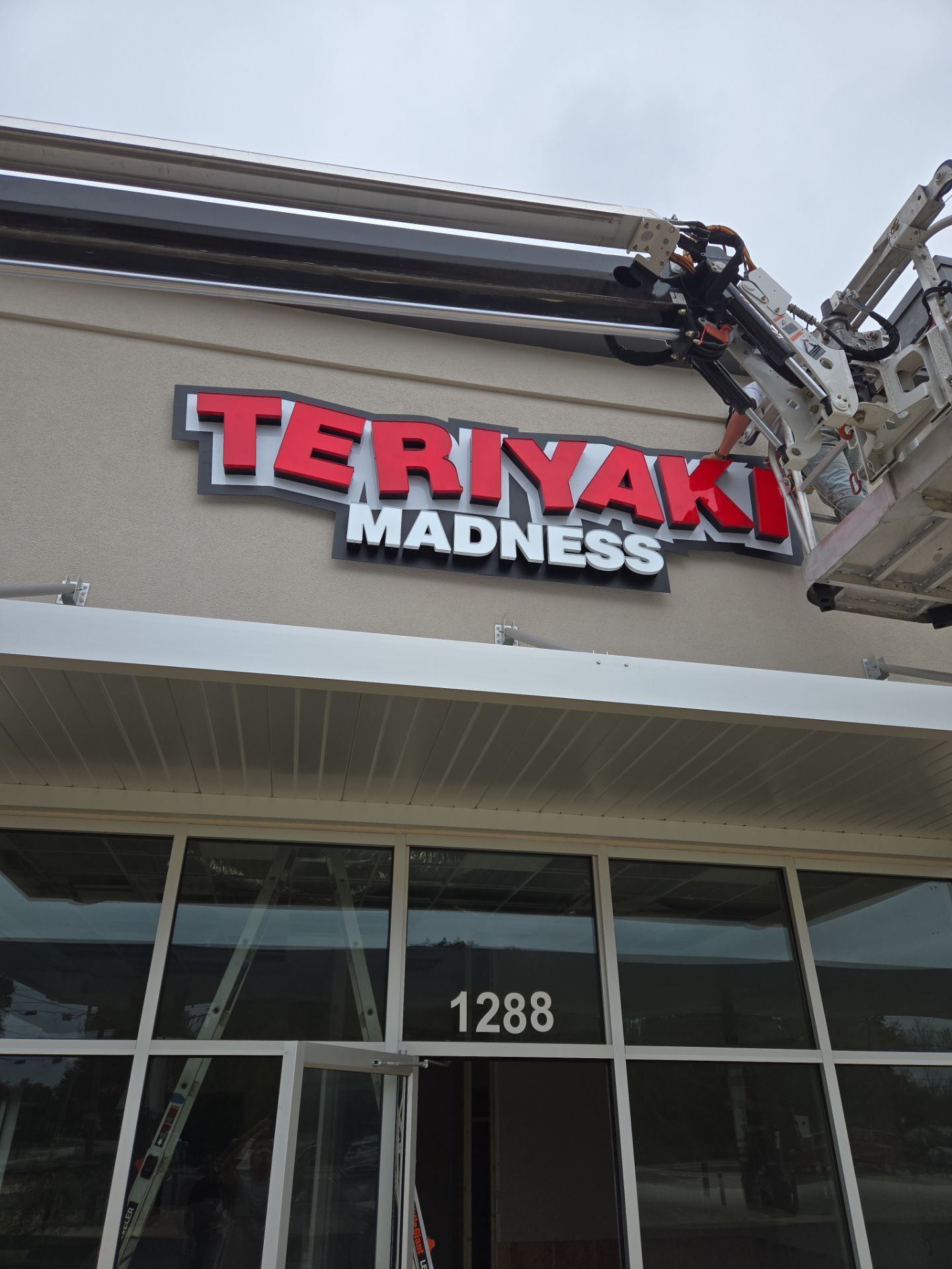 Teriyaki Madness restaurant sign being worked on by a lift. Red and white lettering on a tan building.