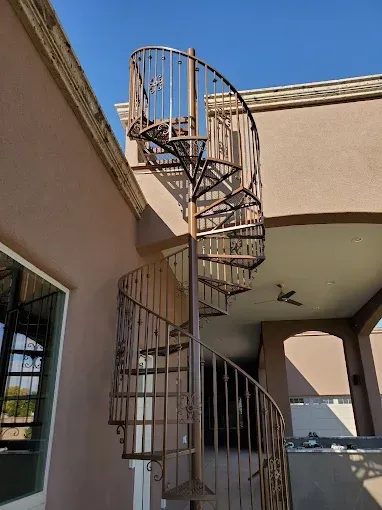 A tall, ornate bronze-colored spiral staircase stands attached to the exterior of a tan-colored stucco building.